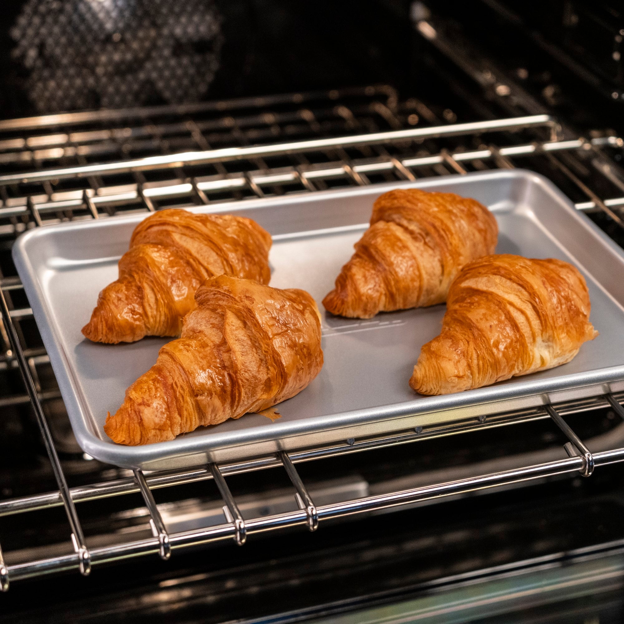 Four golden brown croissants are baking on a silver tray inside an oven. The tray sits on an oven rack, and the croissants look flaky and freshly baked.