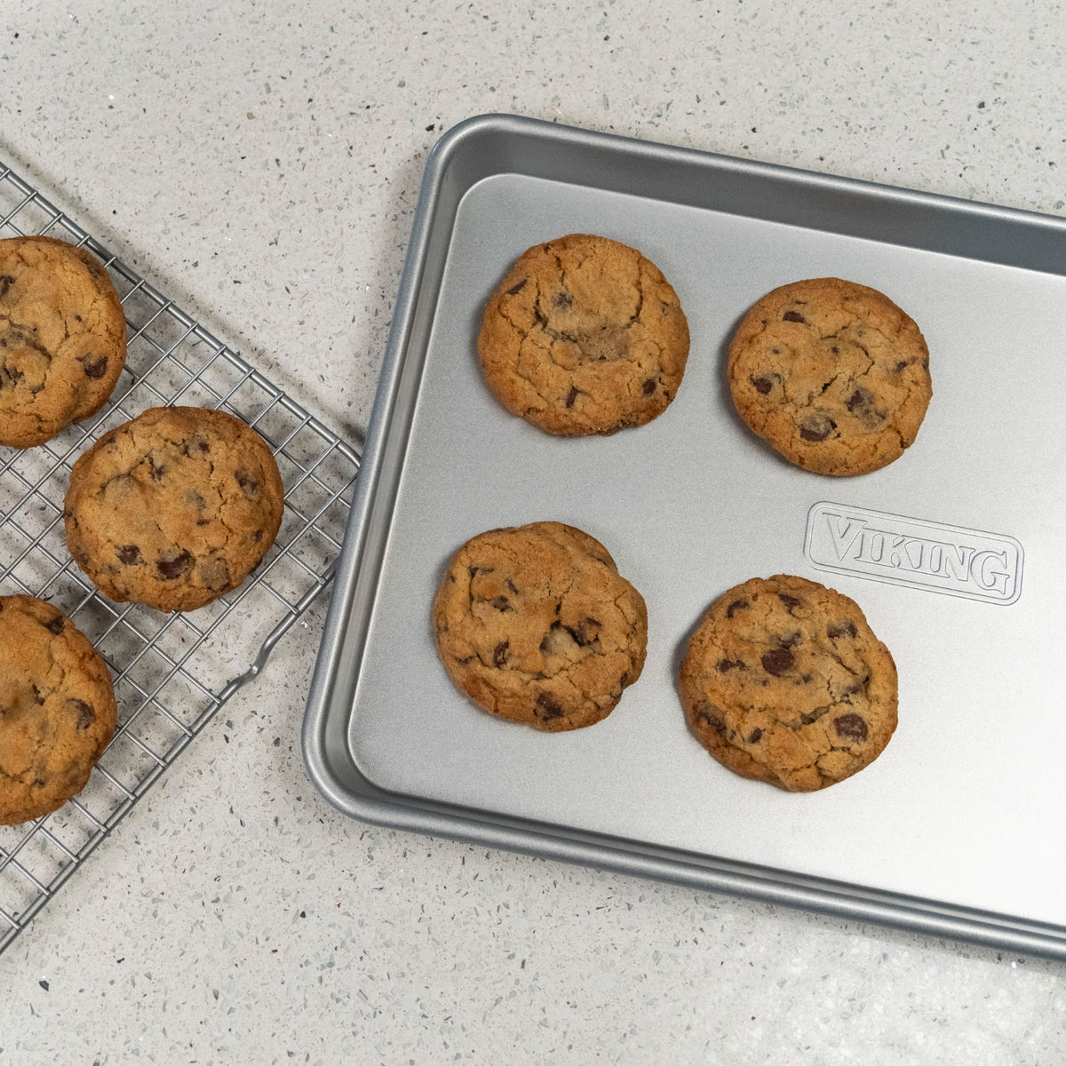 Four chocolate chip cookies on a baking sheet next to five cookies cooling on a wire rack, all on a speckled countertop.