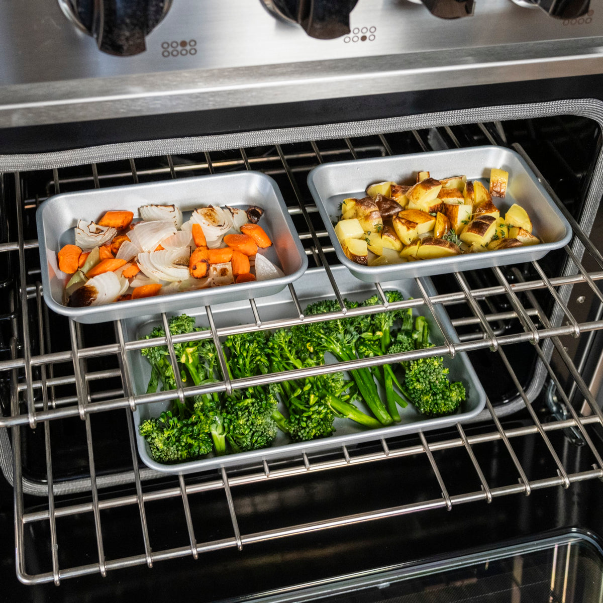 Three baking trays in an oven: one with chopped carrots and onions, one with diced potatoes, and one with broccolini, all being roasted on oven racks.