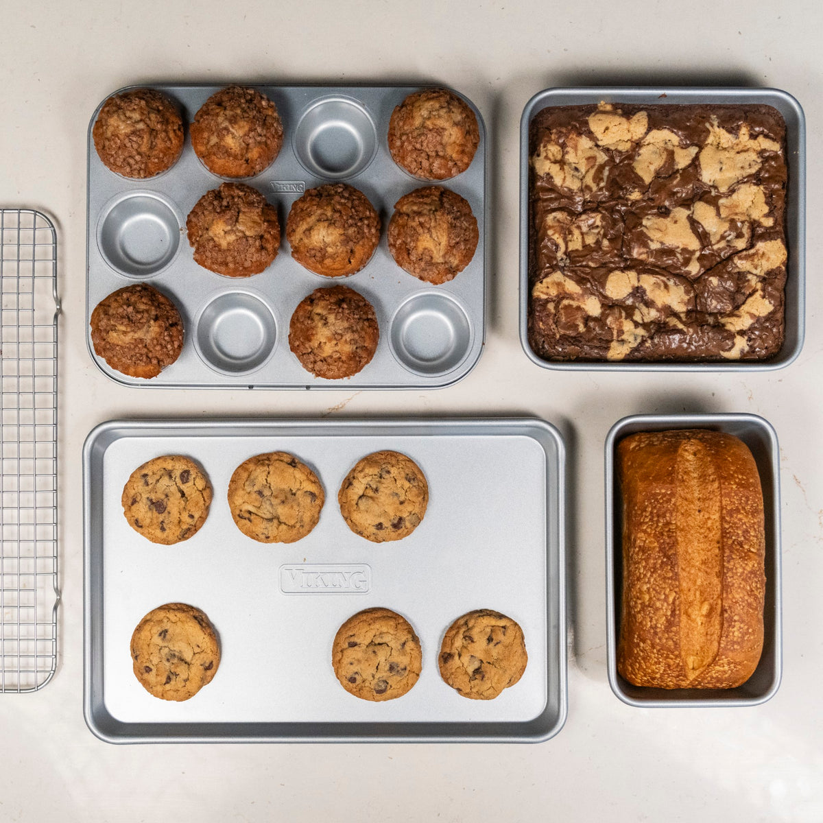 A top-down view of the Viking Aluminized Steel Nonstick 5-pc Bakeware Set by Viking Culinary Products: muffin pan with 12 muffins, square pan of marbled brownies, cooling rack, baking sheet with 6 cookies, and loaf pan with bread.