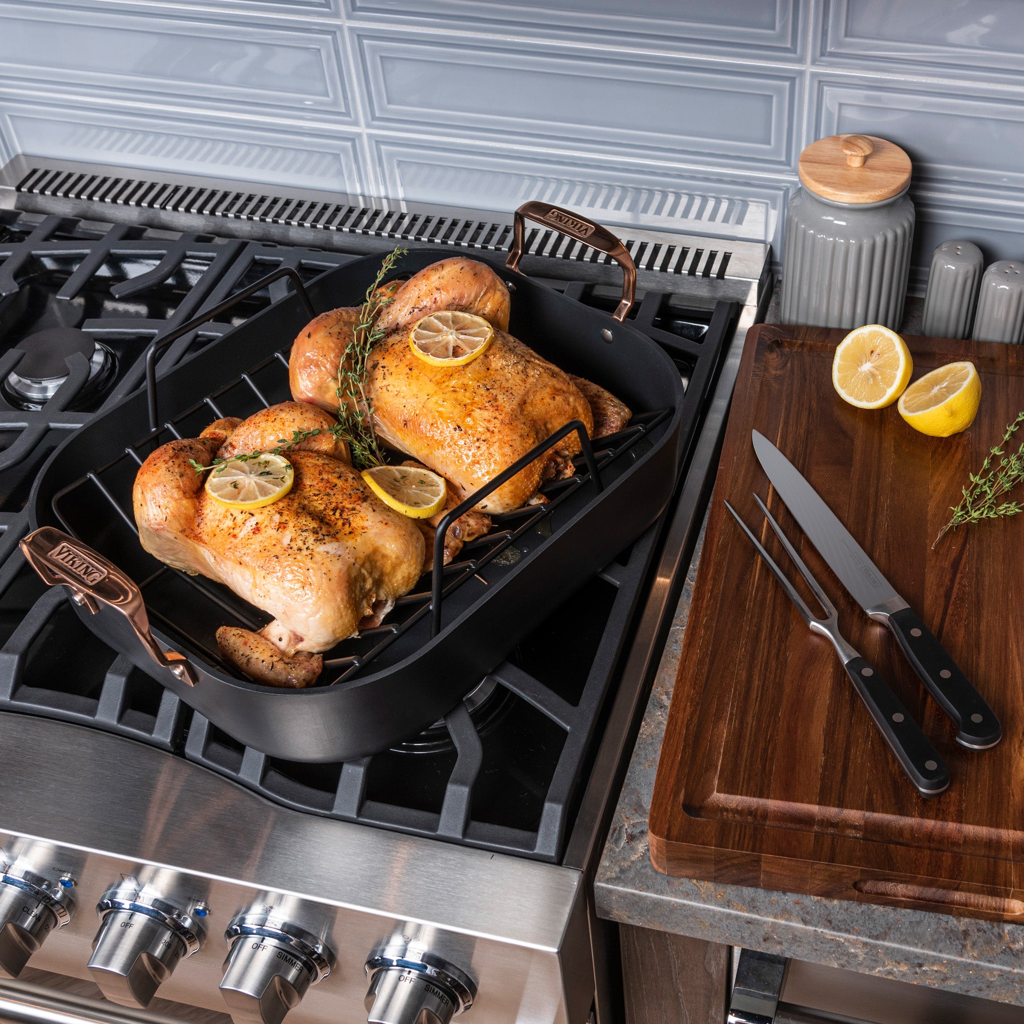 Two roasted chickens with lemon and herbs sit in the Viking Hard Anodized Roaster with Copper Handles from Viking Culinary Products. A carving set and halved lemon rest on a board nearby, with a blue tile backsplash in the background.