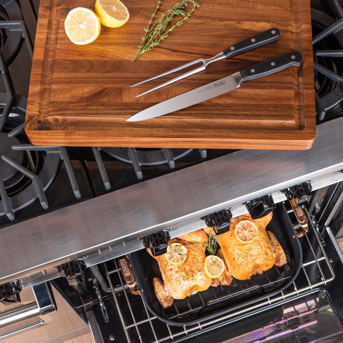A Viking Culinary Products Hard Anodized Roaster with Copper Handles, rack, carving set, and Bluetooth thermometer expertly monitors two chickens roasting in the oven, while herbs and lemon halves rest on a wooden cutting board above.