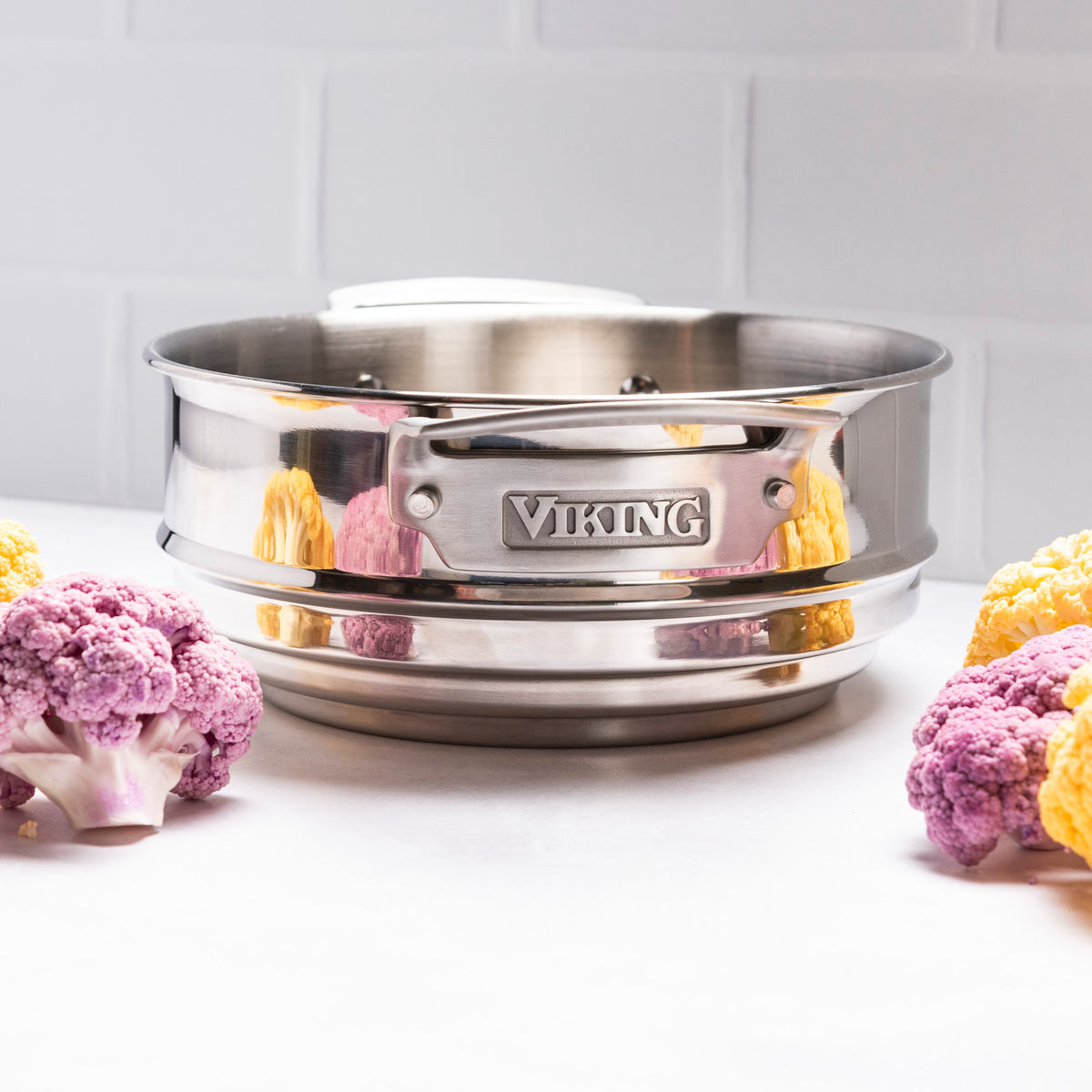 A Viking Culinary Products Hard Anodized Ceramic Nonstick 12pc Cookware set steamer basket rests on a white surface with purple and yellow cauliflower florets, set against a white tiled wall.