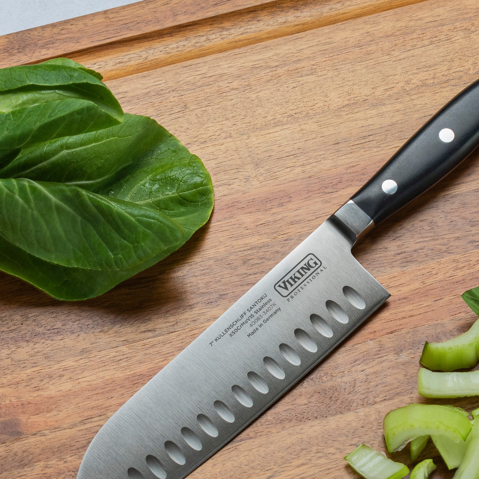 A kitchen knife with a black handle rests on a wooden cutting board, next to fresh lettuce leaves and sliced celery pieces.