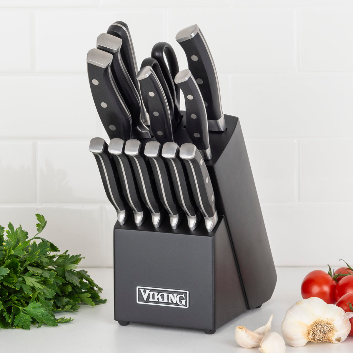 The Viking Culinary Products 14-Piece X50CrMoV15 Steel Cutlery Set with Black Block sits on a countertop beside parsley, garlic, and tomatoes, its sleek forged knives featuring black handles against a white tiled background.