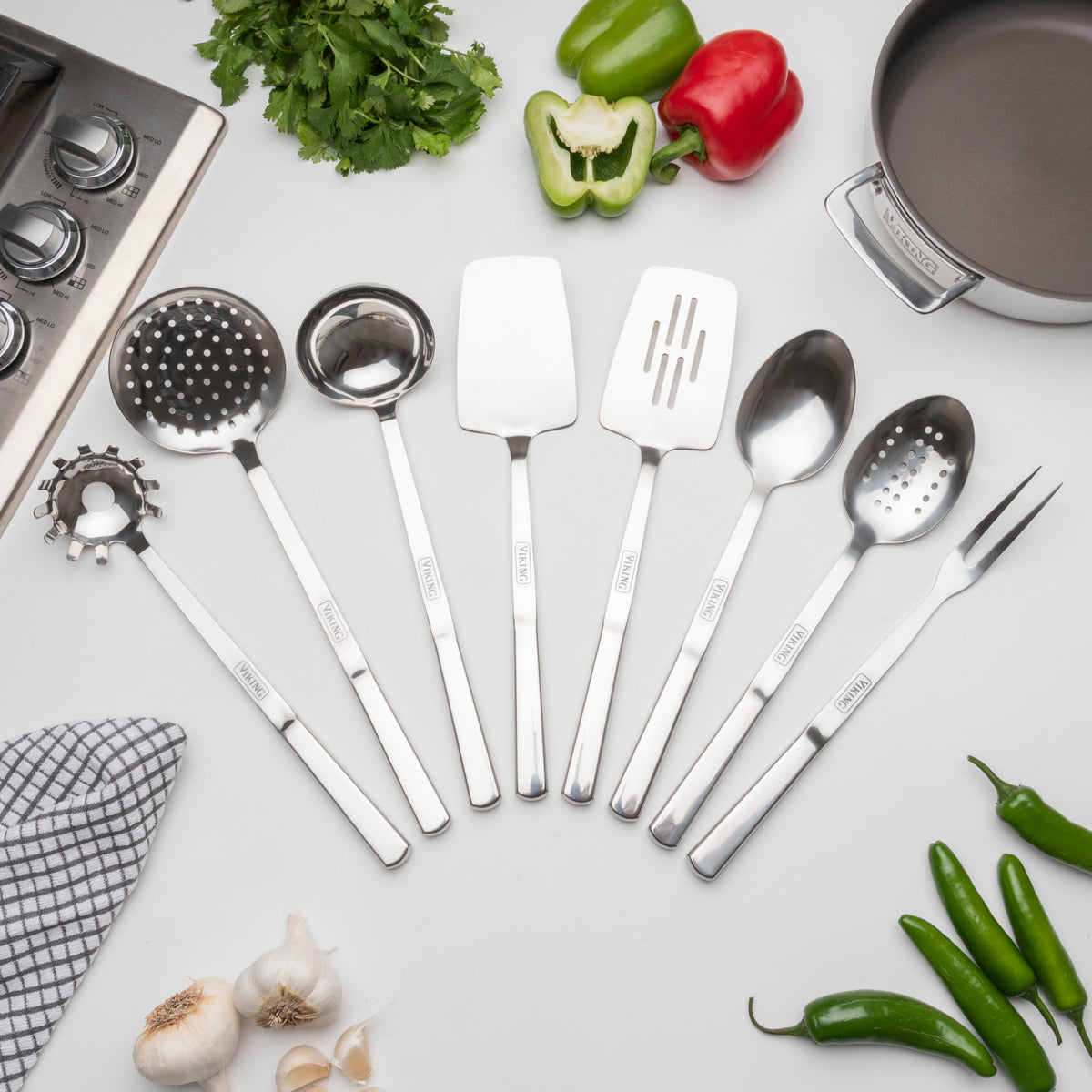 The Viking 8-Piece Stainless Steel Utensil Set from Viking Culinary Products is displayed on a white surface with vegetables and herbs. This durable, dishwasher safe set includes spoons, spatulas, a fork, and a whisk.
