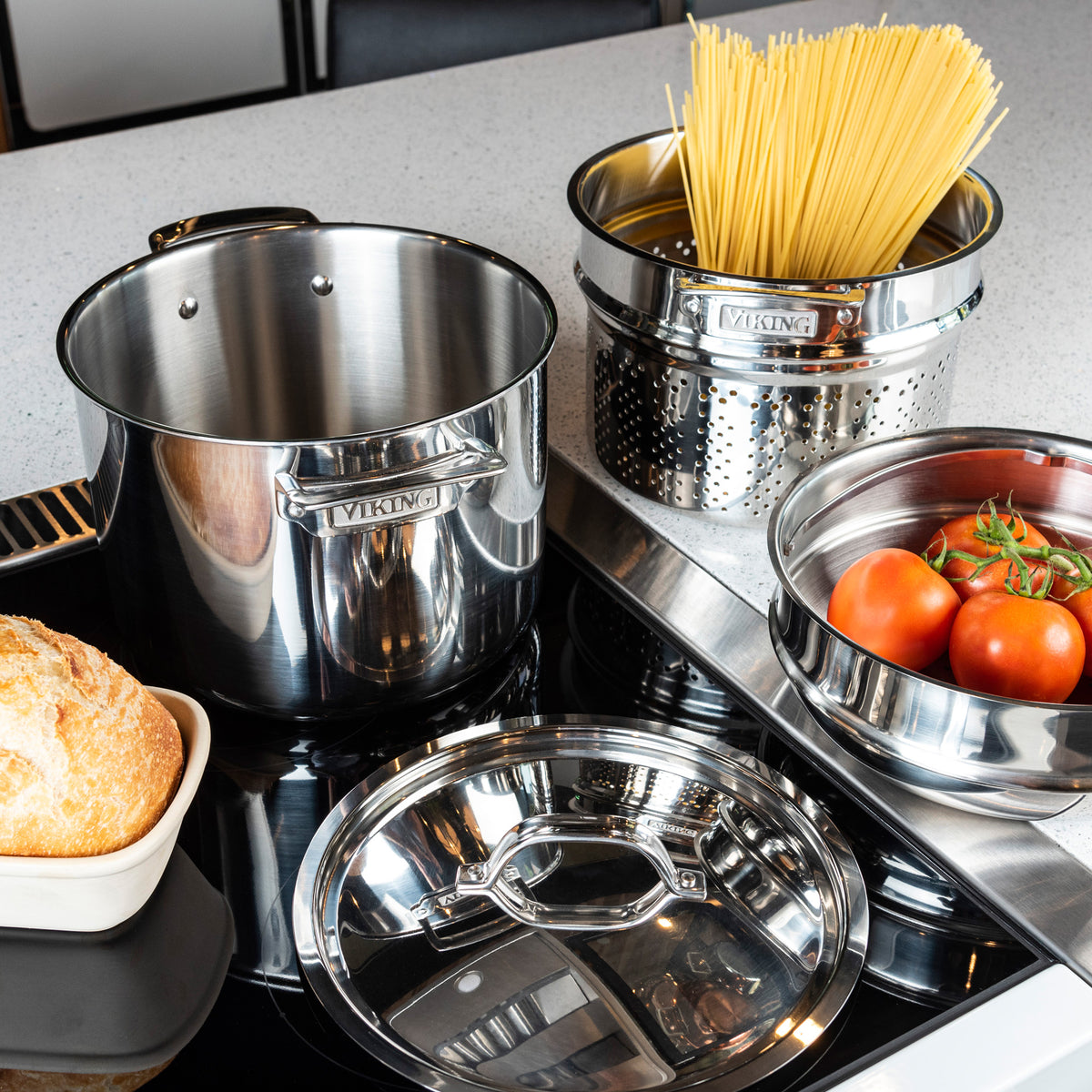 A shiny stainless steel pot set on a stovetop, with uncooked spaghetti in a pasta insert, fresh tomatoes in a bowl, a round loaf of bread in a dish, and a pot lid nearby on a light kitchen counter.