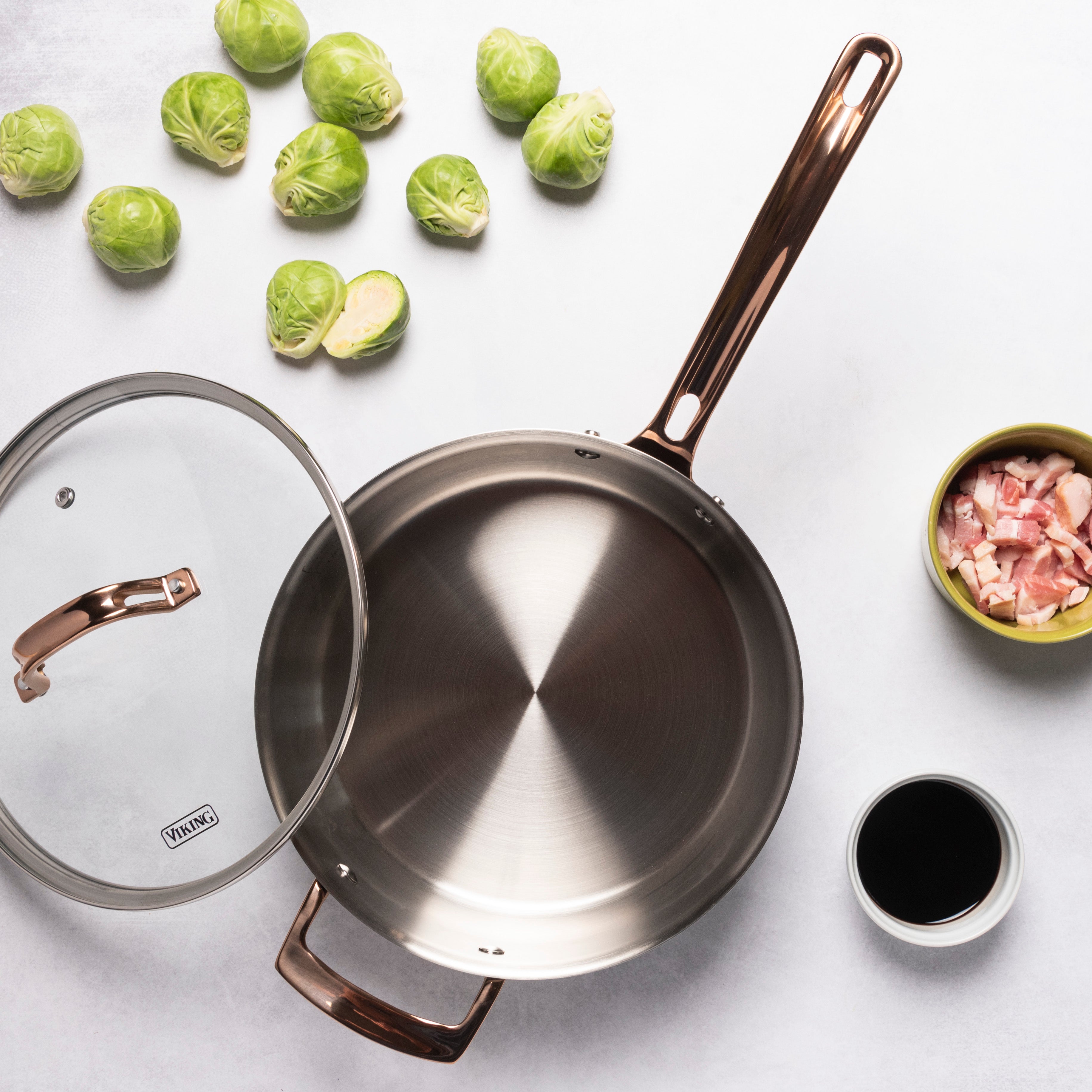 The Viking Culinary Products 3-Ply Black and Copper 4 Quart Sauté Pan with Glass Lid sits nearby, surrounded by Brussels sprouts, chopped bacon, and a dish of dark liquid on a light surface. Induction compatible for versatile cooking.