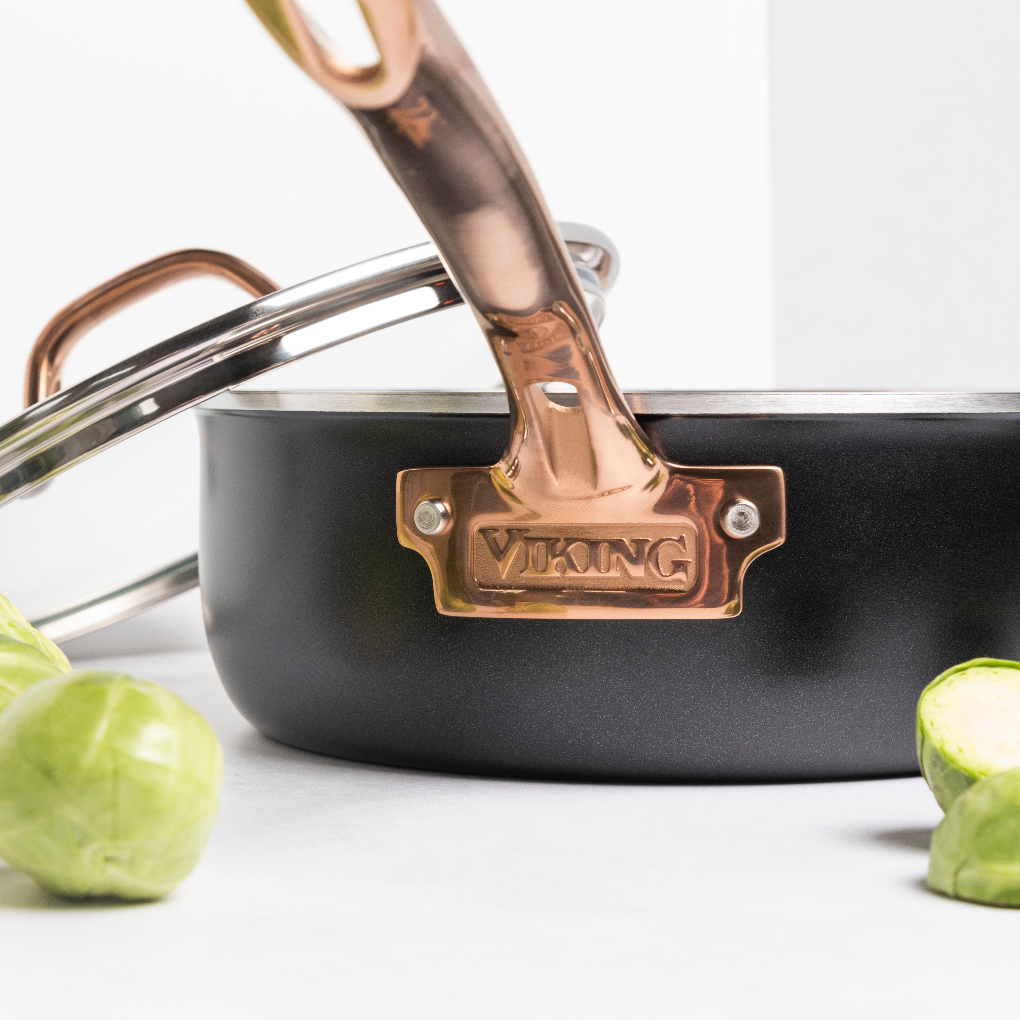 A close-up of the Viking Culinary Products 3-Ply Black and Copper 4 Quart Sauté Pan with Glass Lid, surrounded by fresh Brussels sprouts on a white surface; the induction compatible lid is partially visible.