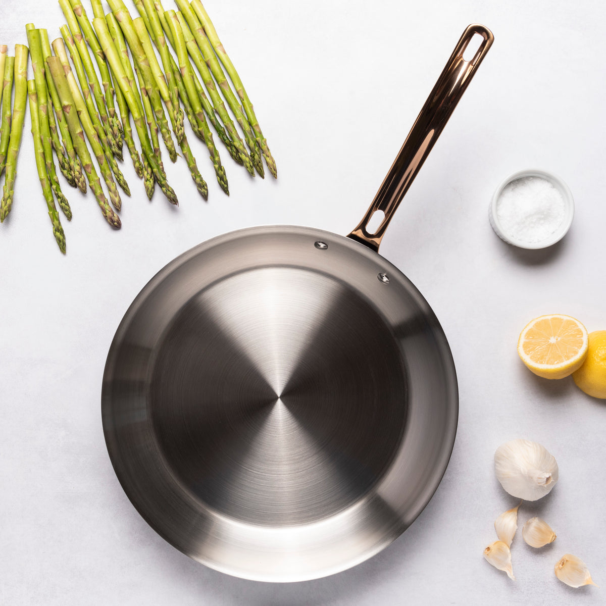 The Viking 3-Ply Black and Copper 12 Inch Fry Pan by Viking Culinary Products rests on a light surface, surrounded by fresh asparagus, a bowl of salt, two lemons (one sliced), and several garlic cloves.