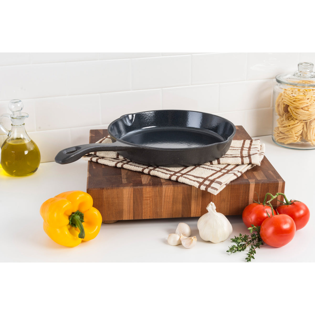 A black cast iron skillet sits on a folded kitchen towel atop a wooden cutting board, surrounded by a yellow bell pepper, garlic, tomatoes, herbs, a jar of pasta, and a bottle of olive oil.