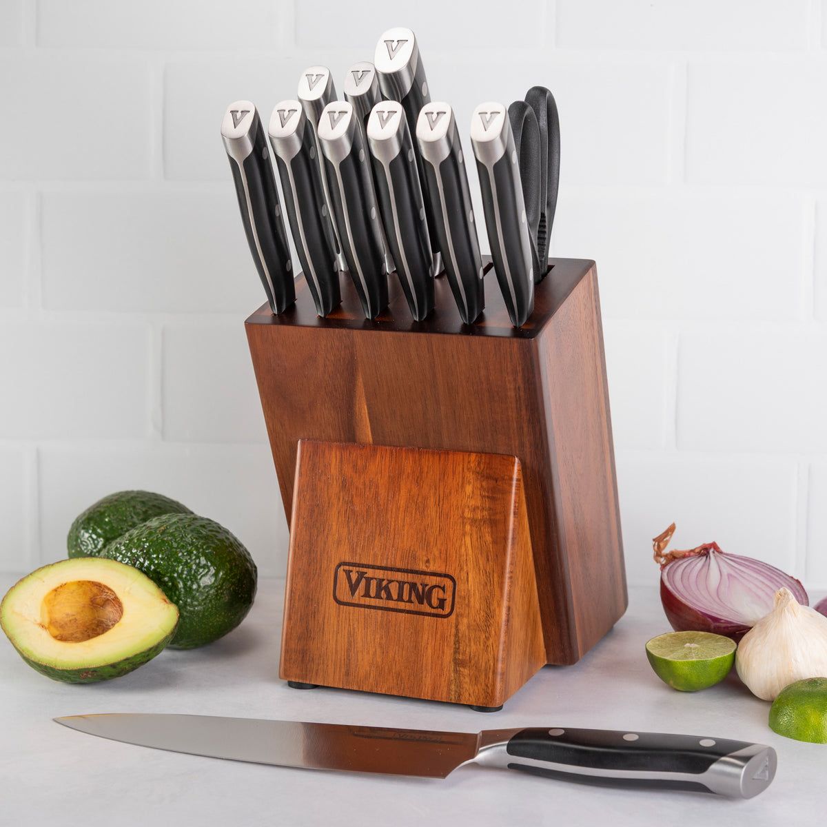 The Viking 12-Piece Stainless Steel Forged Cutlery Block Set by Viking Culinary Products is displayed on a counter with a chef’s knife, halved avocado, lime, and shallot against a white tiled background.