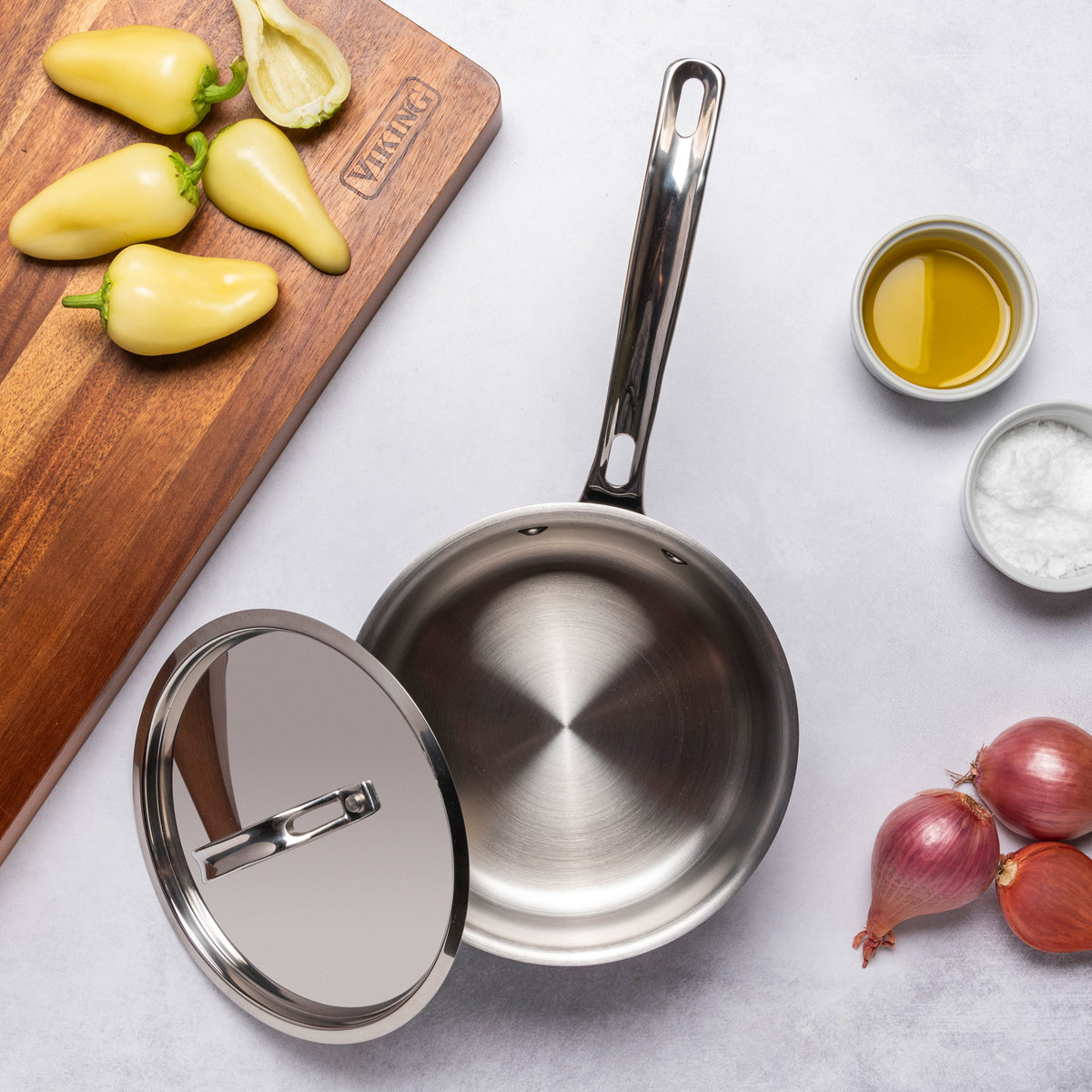 A PFAS-free Viking 12pc 3-Ply Cast Black Exterior Stainless Steel saucepan with a metal lid is on a light surface near yellow peppers on a wooden board, olive oil, salt, and two shallots. Induction compatible. Brand: Viking Culinary Products.