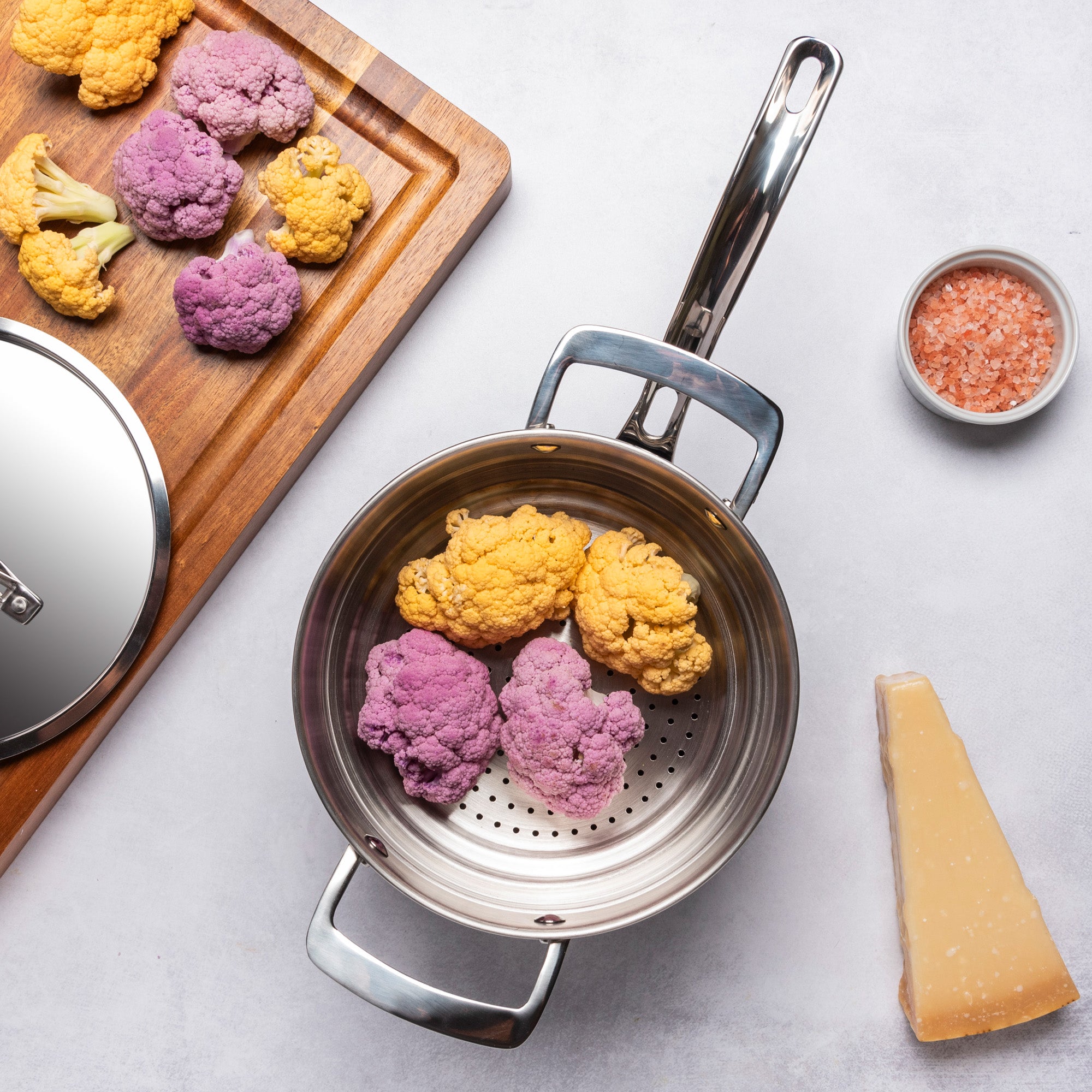 A PFAS-free Viking Culinary Products 12pc 3-Ply Cast Black Stainless Steel steamer pot with metal lid sits on a light surface, filled with purple and yellow cauliflower, alongside cheese, pink salt, and more cauliflower on a wooden board.