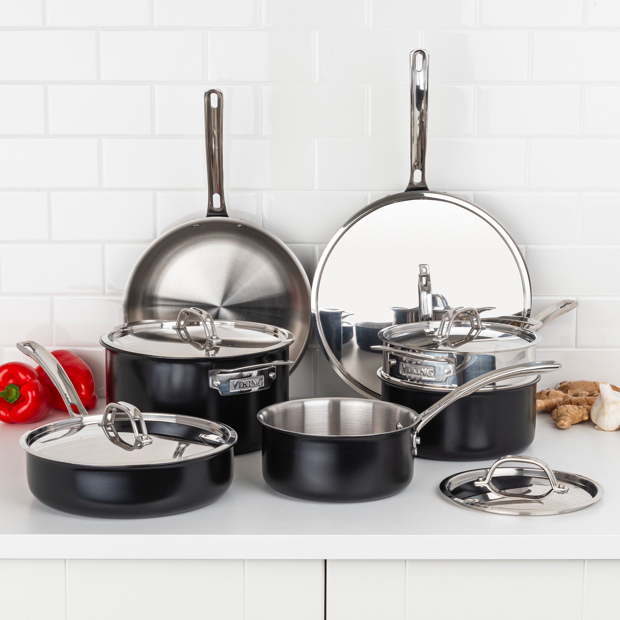 The Viking 12pc 3-Ply Cast Black Exterior Stainless Steel Cookware Set by Viking Culinary Products with metal lids is displayed on a white kitchen counter, surrounded by red peppers, ginger, and garlic.