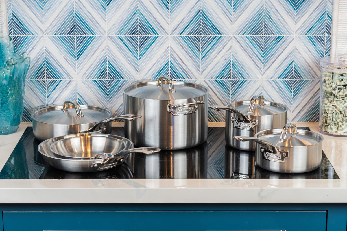 A set of stainless steel pots and pans with lids sits on a stovetop, with a blue and white geometric backsplash in the background. The cookware is arranged neatly, reflecting the kitchens modern and clean style.