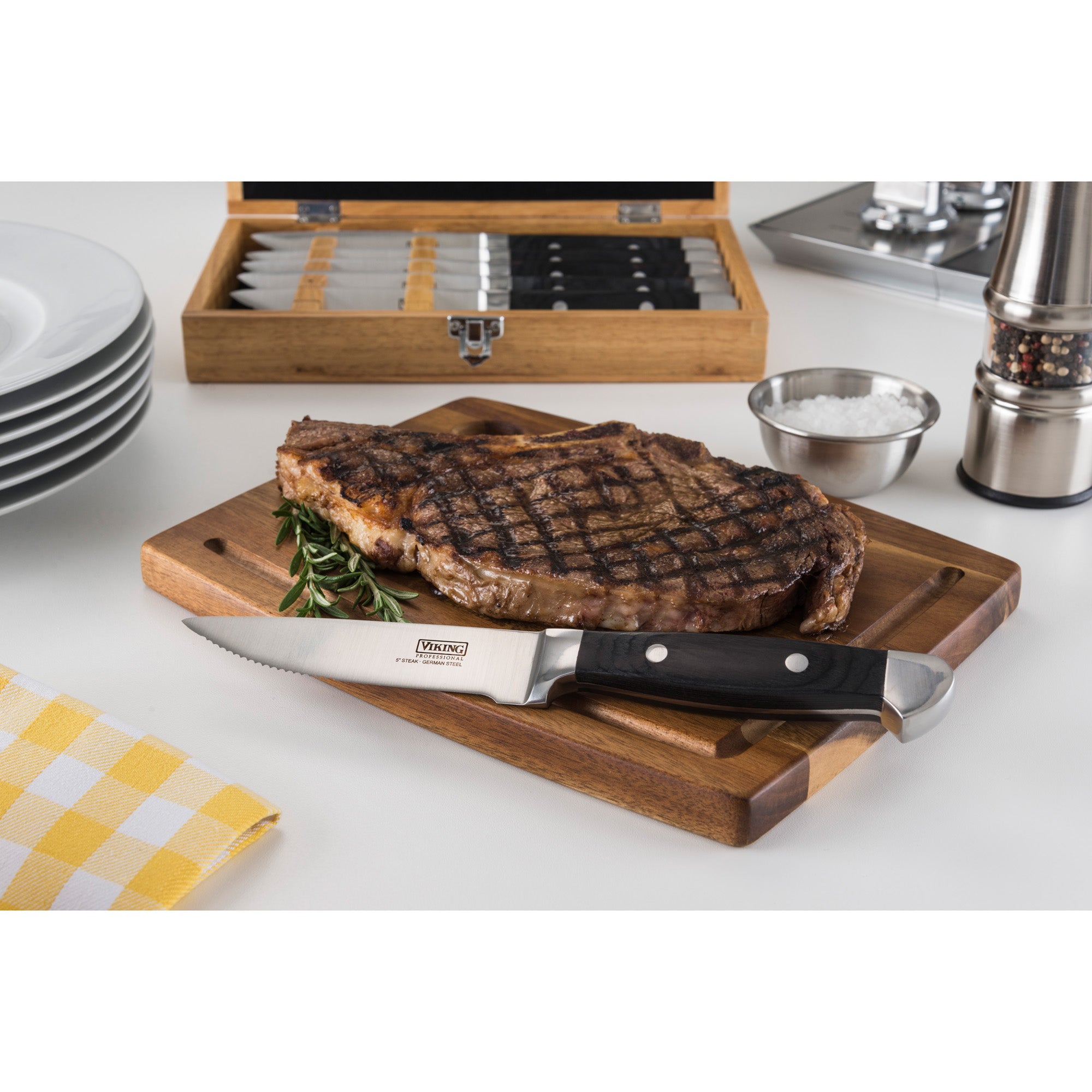 A grilled steak rests on a wooden cutting board with a steak knife in front. In the background are a box of knives, a stack of plates, a metal spice shaker, a small bowl, and a yellow checked napkin.