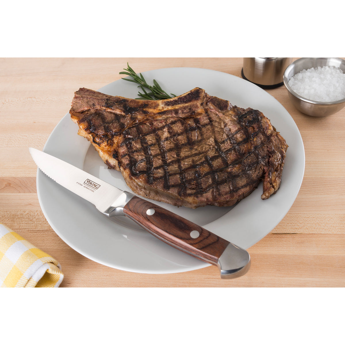A grilled steak with visible grill marks sits on a white plate, accompanied by a steak knife with a wooden handle. The plate is on a wooden surface, with a sprig of rosemary and a bowl of coarse salt nearby.