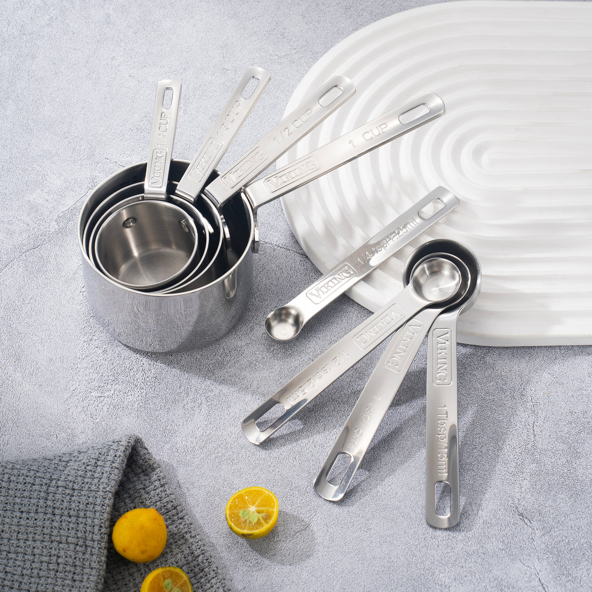 The Viking Stainless Steel Measuring Cup Set by Viking Culinary Products is arranged on a gray surface with a white tray, gray cloth, and halved lemons. These durable measuring cups are dishwasher safe for easy cleaning.