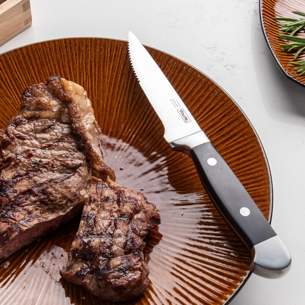 A grilled steak is served on a brown plate with a serrated steak knife placed beside it. The background includes a partial view of a plate with herbs.