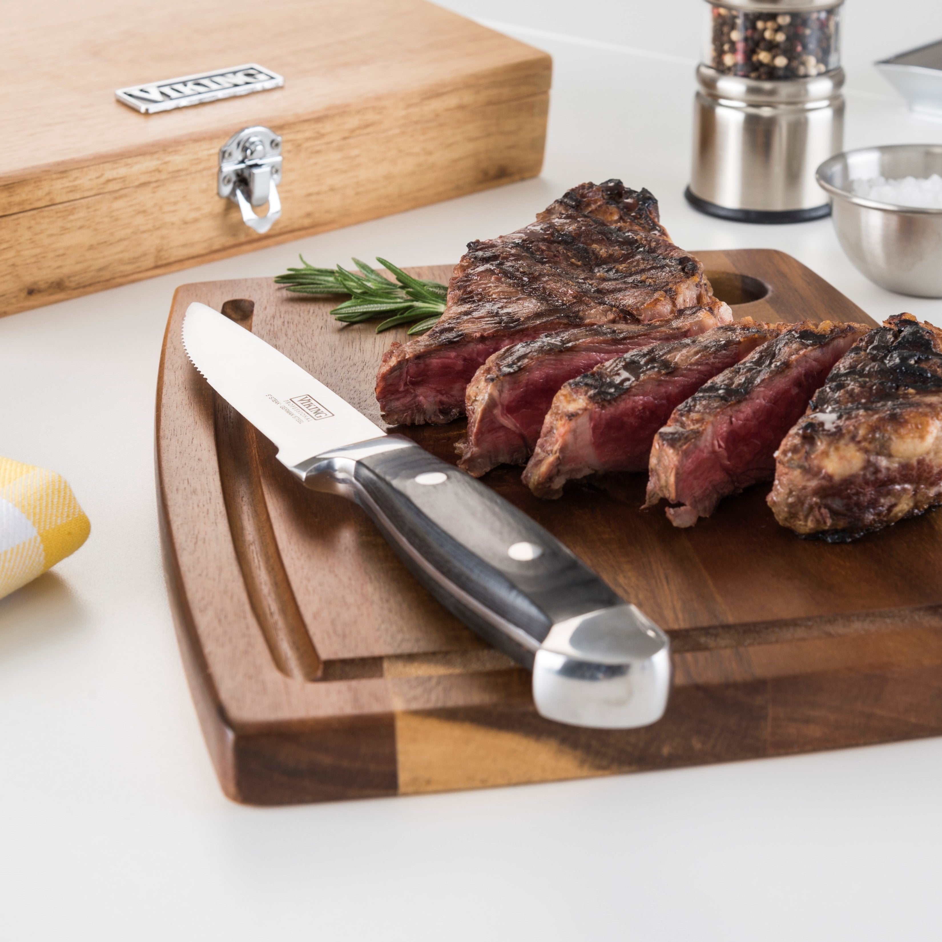 A grilled steak sliced on a wooden cutting board with a steak knife, rosemary garnish, and a wooden case, pepper grinder, and small bowl of salt in the background.