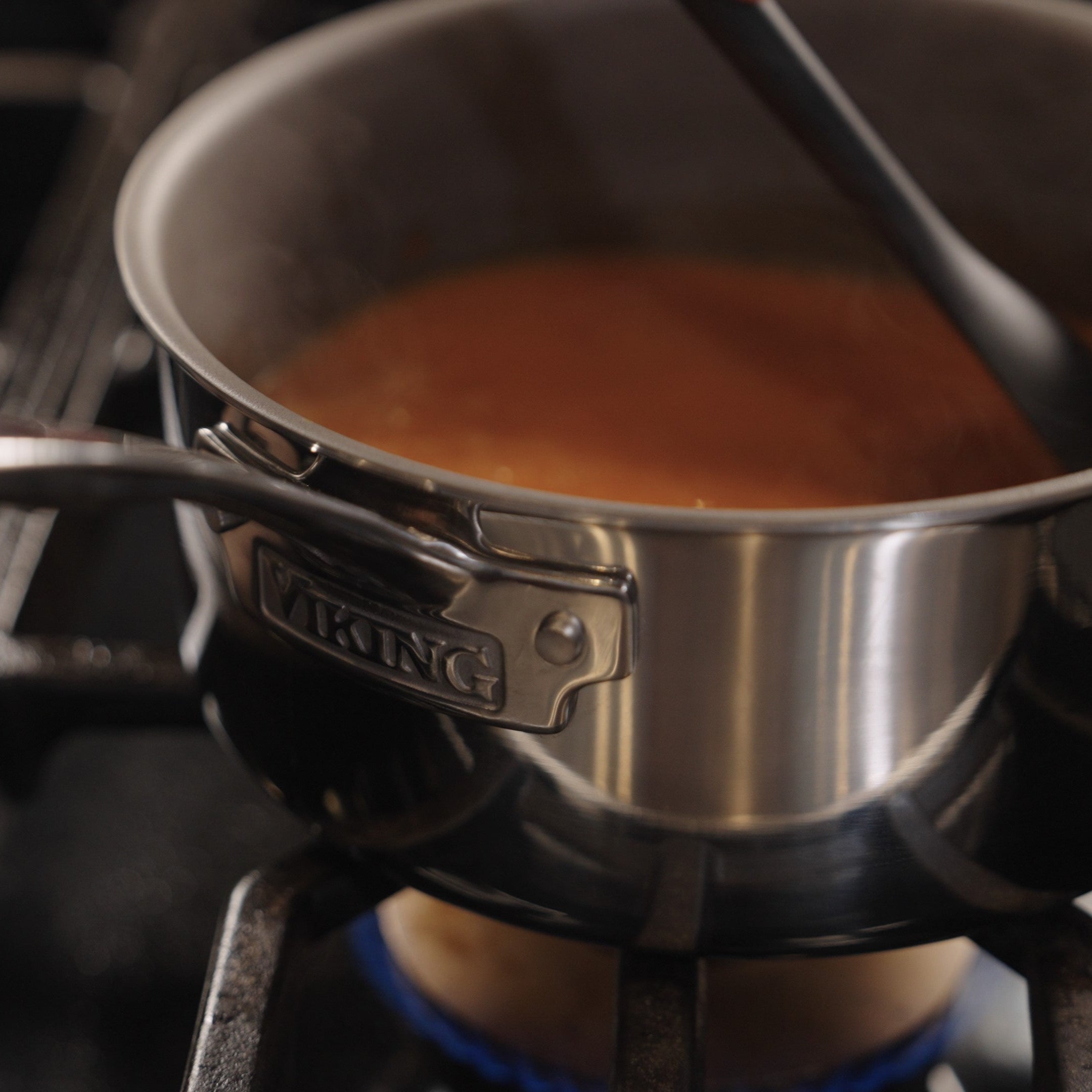 The Viking PureGlide Pro 3-Ply 3-Quart Sauce Pan by Viking Culinary Products simmers a brown liquid on a gas stove, steam rising as a utensil stirs in the chemical-free nonstick cookware with its metal lid.
