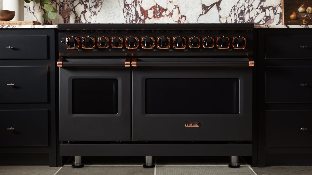 A black double oven range with copper accents and multiple knobs, set in a modern kitchen with dark cabinets and a decorative marble backsplash.