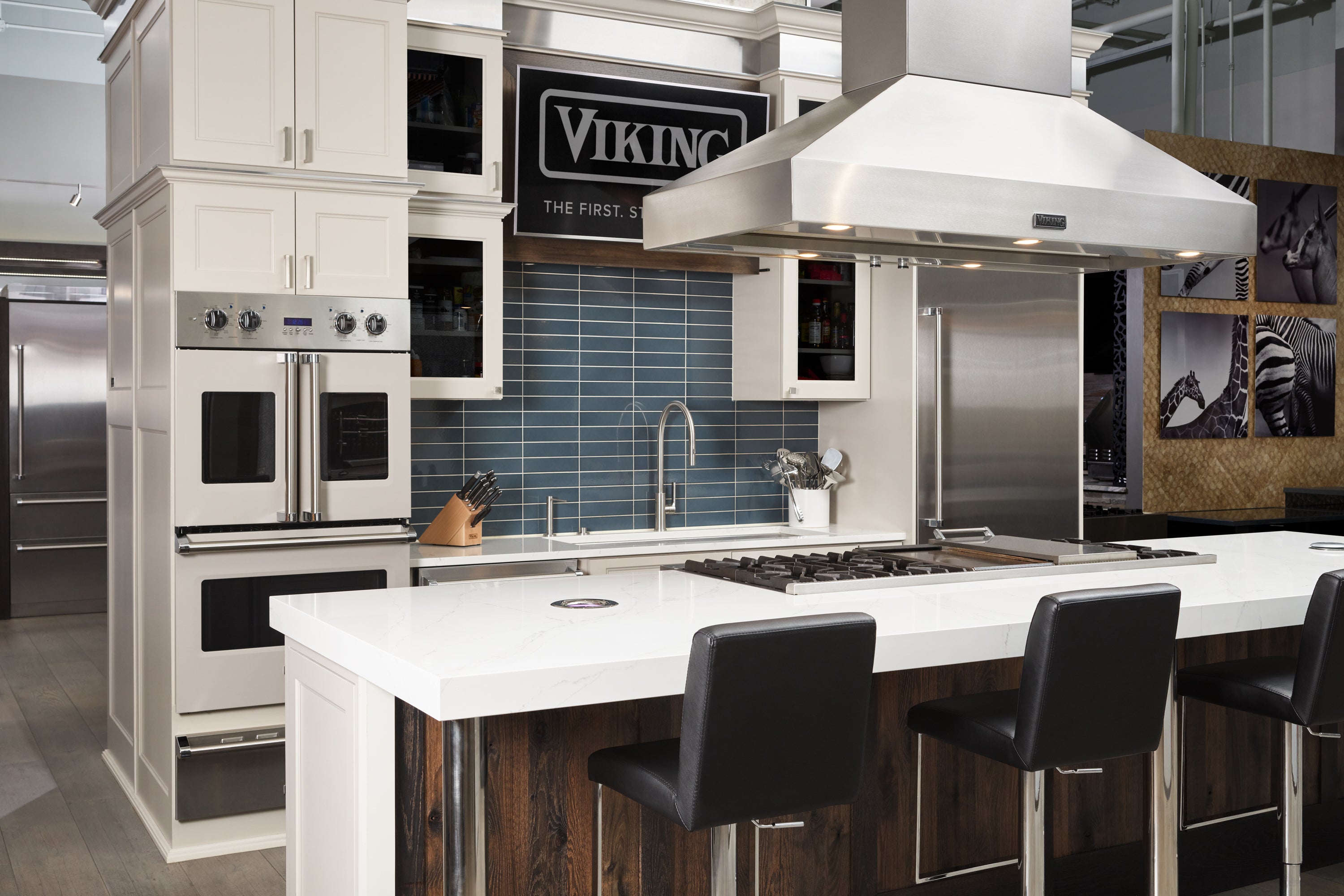 Modern kitchen with white cabinets, blue tile backsplash, stainless steel appliances, a large island with a stove and range hood, and three black barstools.