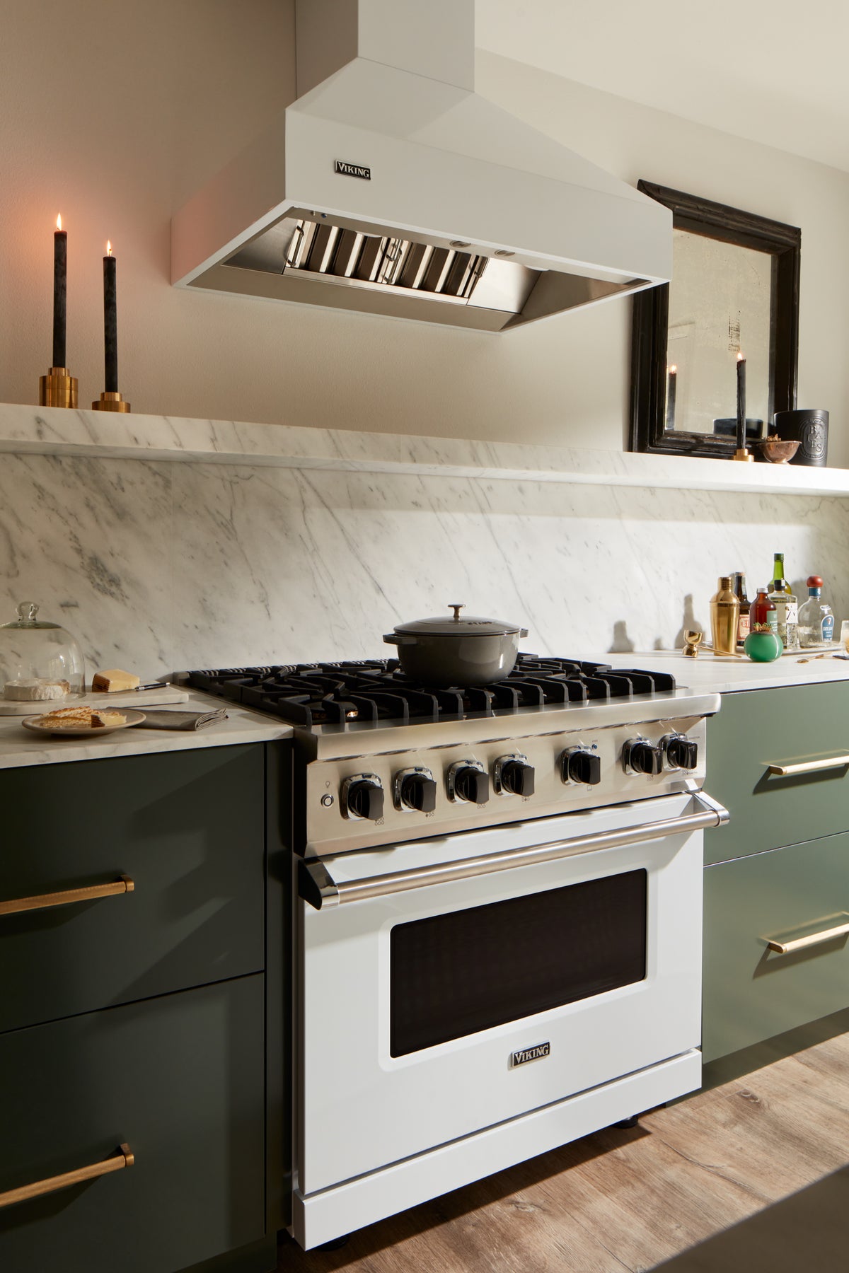 A modern kitchen with a white Maytag gas stove and oven, green cabinets with gold handles, marble backsplash, two lit black candles, and various kitchen items on the countertop.
