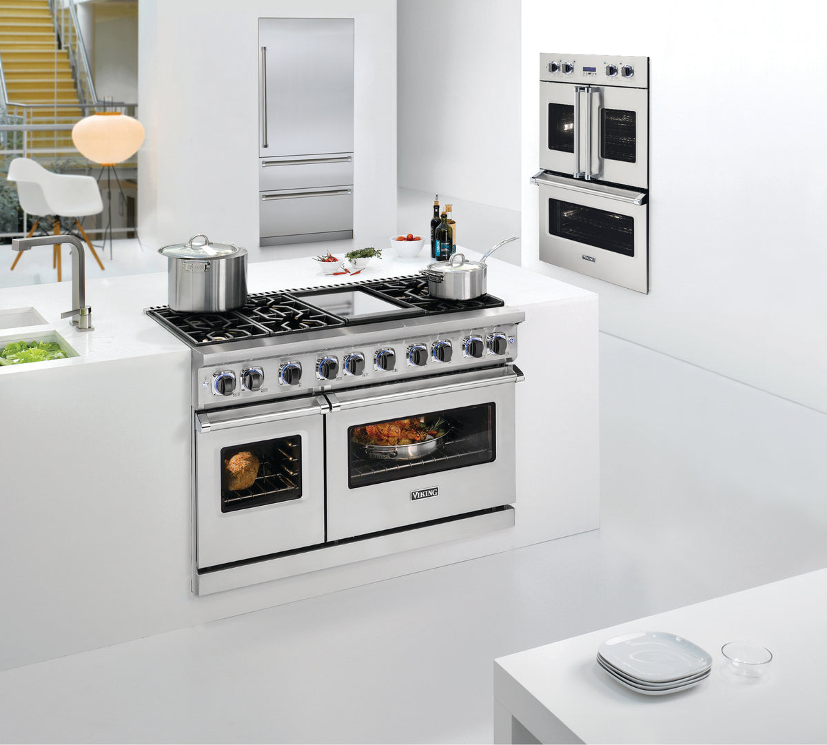 Modern, sleek white kitchen with a stainless steel stove featuring double ovens, two pots on the stovetop, a built-in wall oven, and minimalistic decor. Plates are set on a white counter in the foreground.