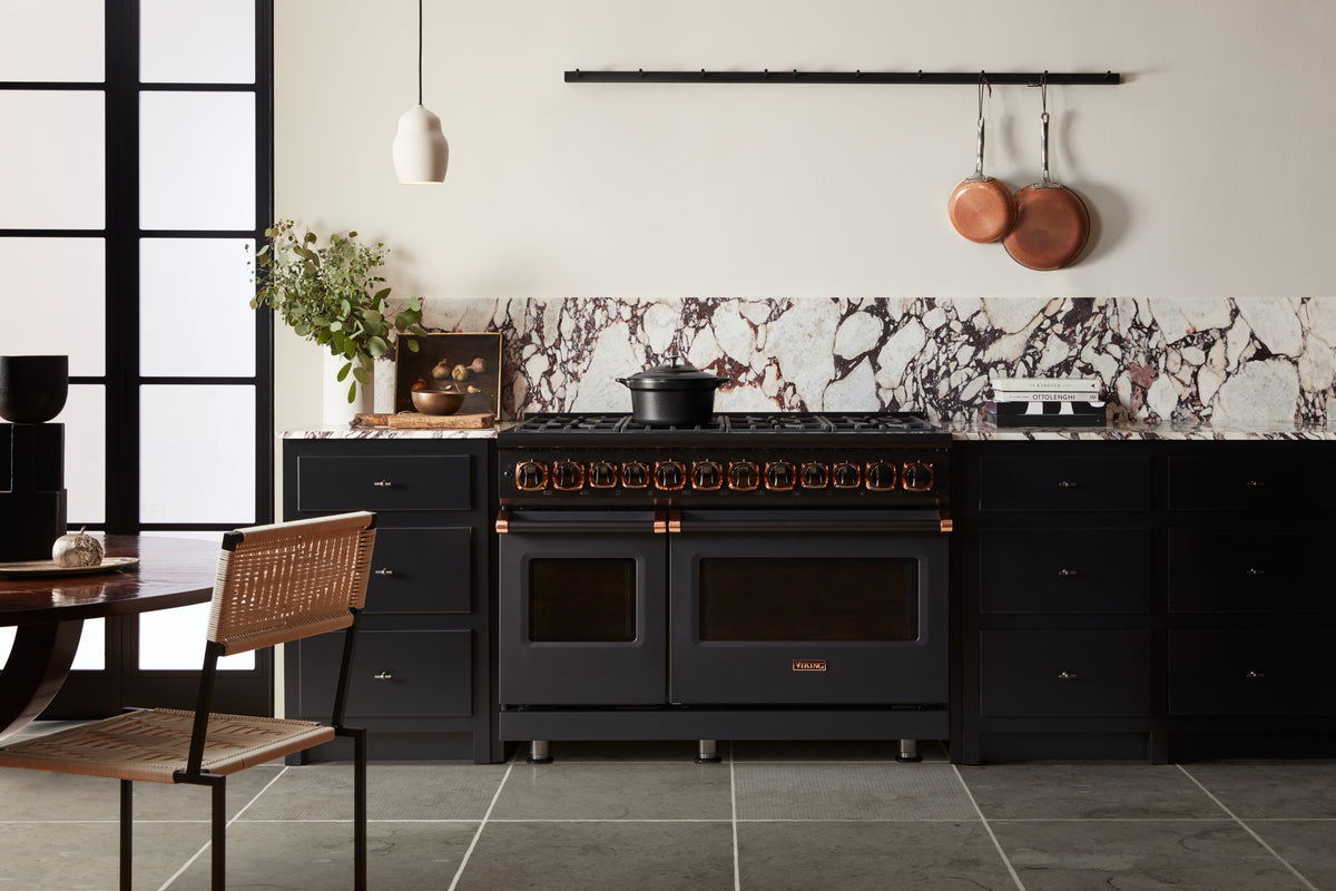 A modern kitchen with black cabinets, a large black stove, marble backsplash, hanging copper pans, a plant, and a small dining table with a wicker chair. The space is sleek and minimalist with neutral tones.