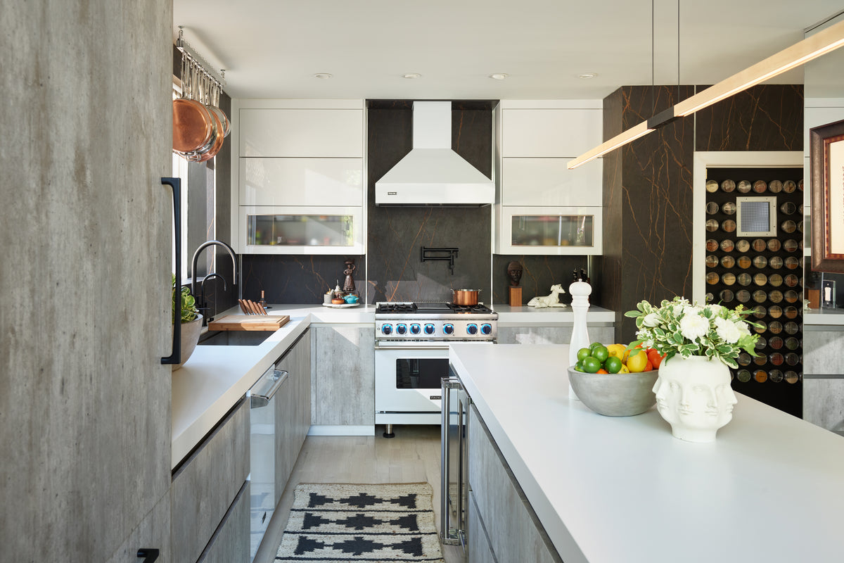 Modern kitchen with white countertops, stainless steel stove, black backsplash, an island with a vase of flowers and fruit, a wall-mounted spice rack, and minimalist decor.