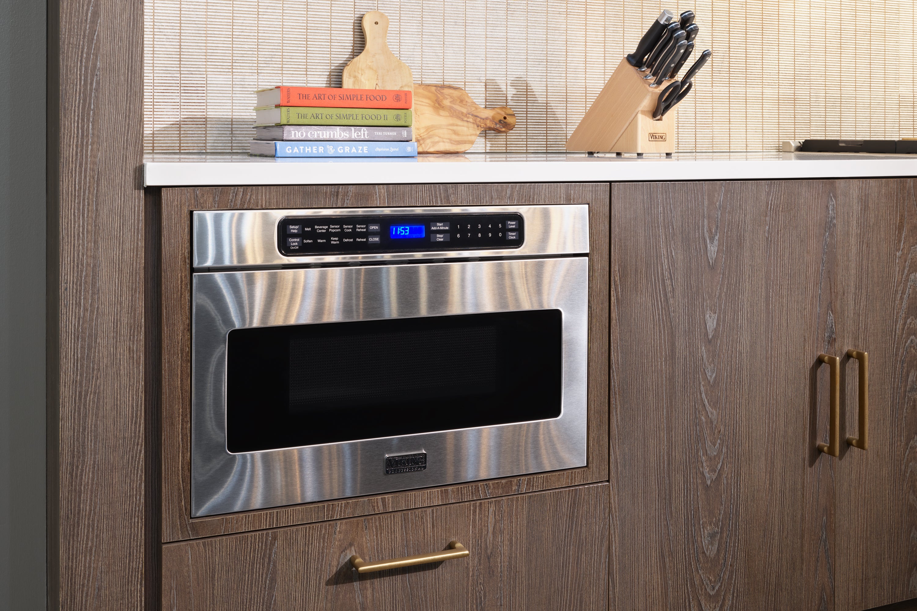 A stainless steel microwave built into a wood kitchen cabinet under a white countertop. On the counter are cookbooks, a cutting board, a loaf of bread, and a knife block with knives.