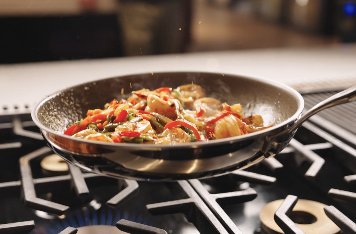 A stainless steel skillet on a gas stove contains sautéed vegetables and shrimp, including bell peppers and onions, being tossed in mid-air while cooking.