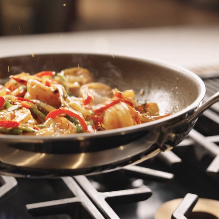 A stainless steel pan on a stovetop filled with shrimp, sliced red bell peppers, and green beans being sautéed, with some food slightly airborne as it is tossed in the pan.