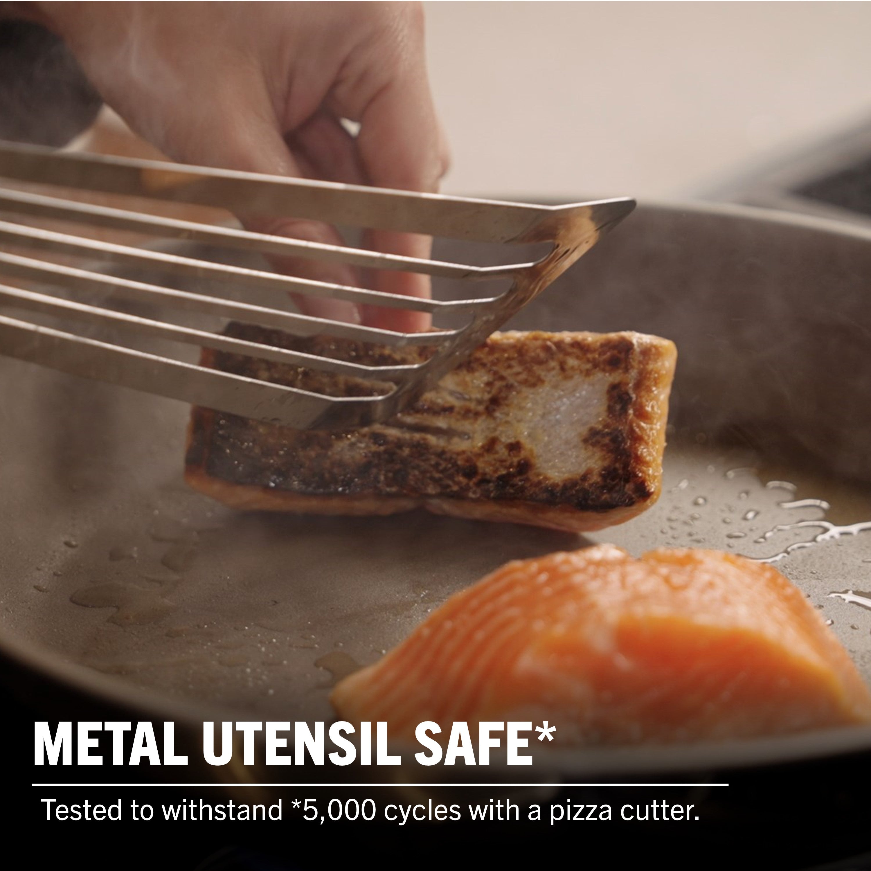A hand uses a metal spatula to lift cooked fish from the Viking PureGlide Pro 3-Ply Fry Pan, with raw salmon beside it. Text: METAL UTENSIL SAFE*—withstands 5,000 cycles with a pizza cutter. Viking Culinary Products.