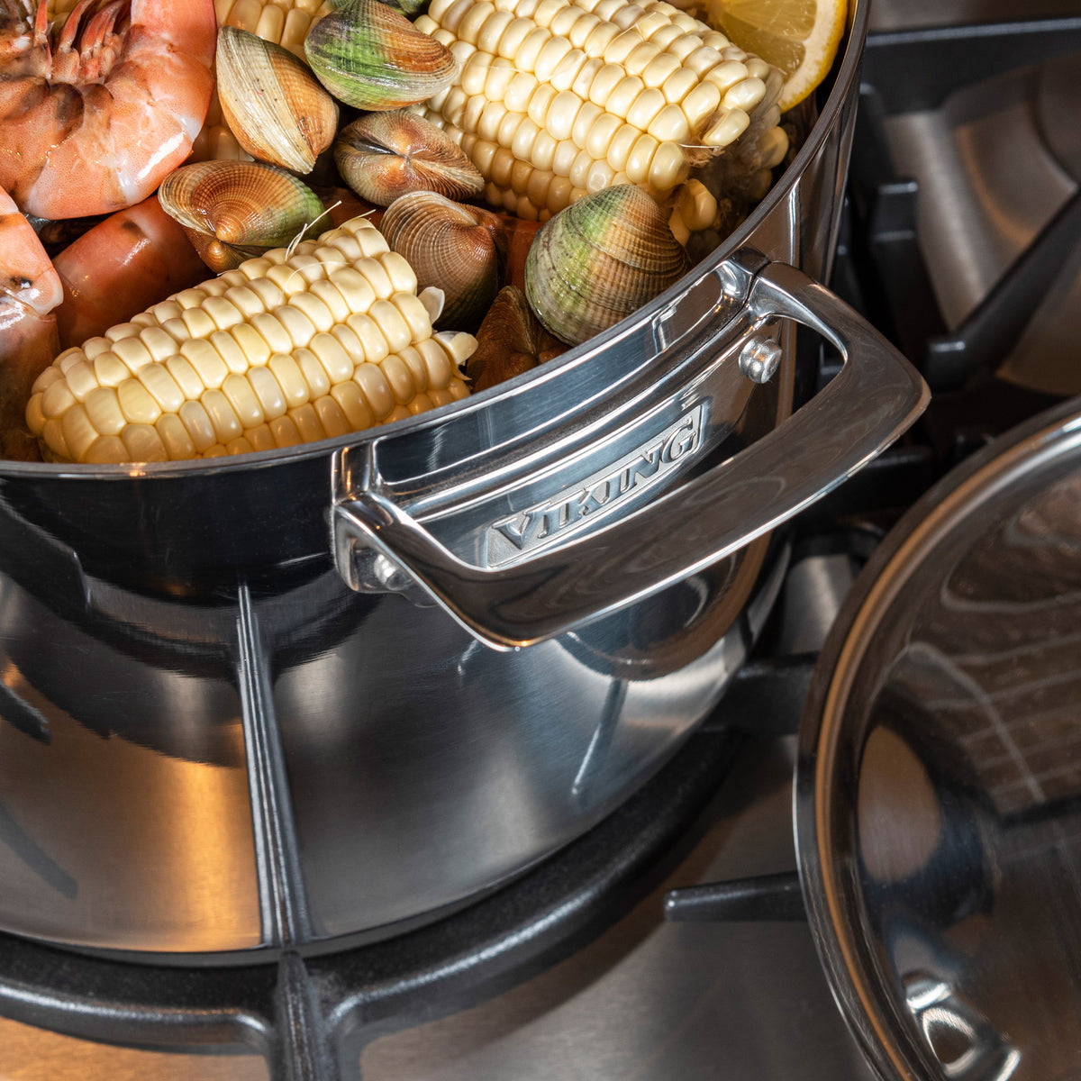 The Viking Culinary Products 3-Ply Stainless Steel 12-Quart Stock Pot with Metal Lid sits on the stove, filled with corn, clams, and shrimp ready to boil. Its induction capable and dishwasher safe for easy cleanup.