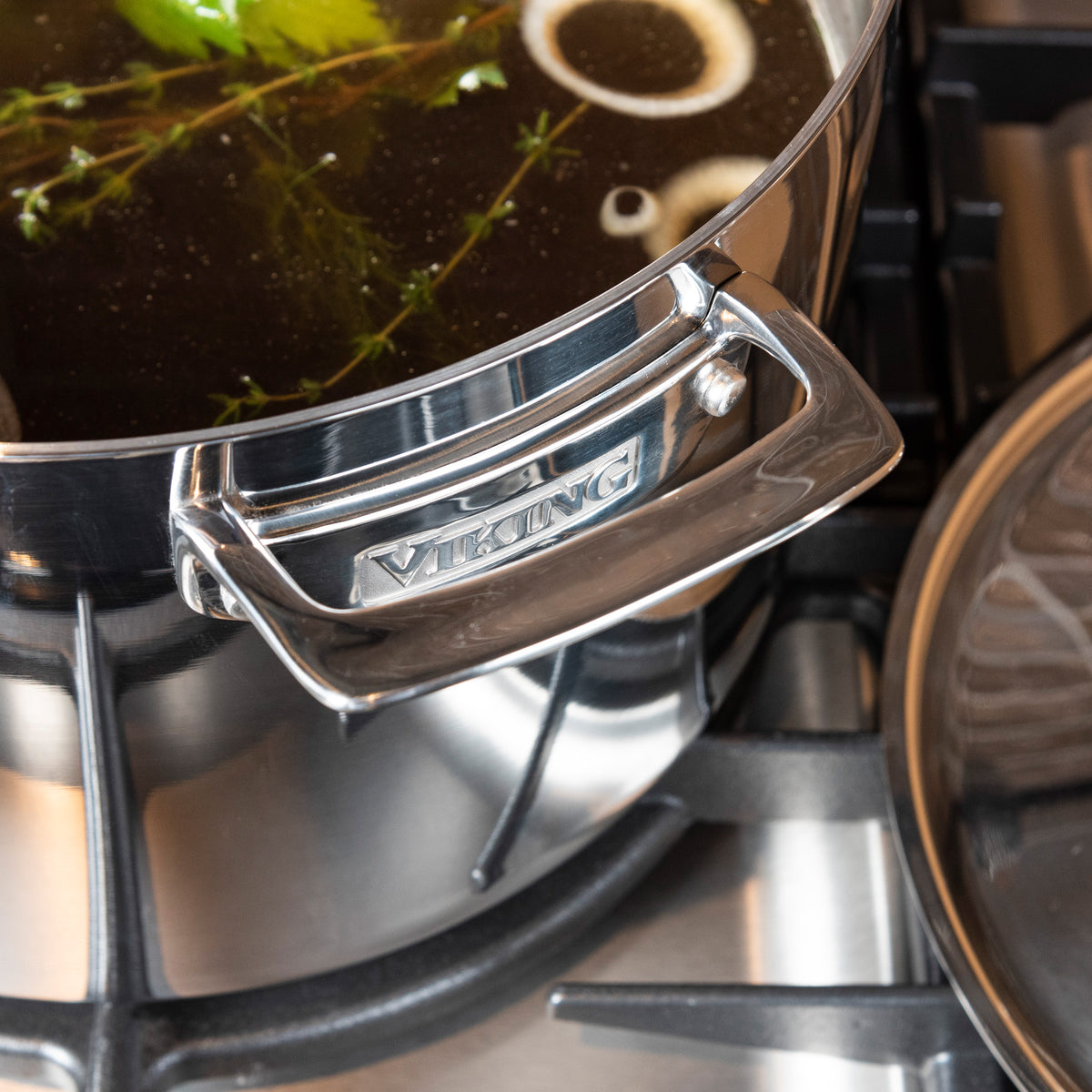 A close-up of the Viking Culinary Products 3-Ply Stainless Steel 12-Quart Stock Pot with Metal Lid on a stove, filled with broth, herbs, and onions. The induction-capable handle and logo highlight its premium stainless steel design.