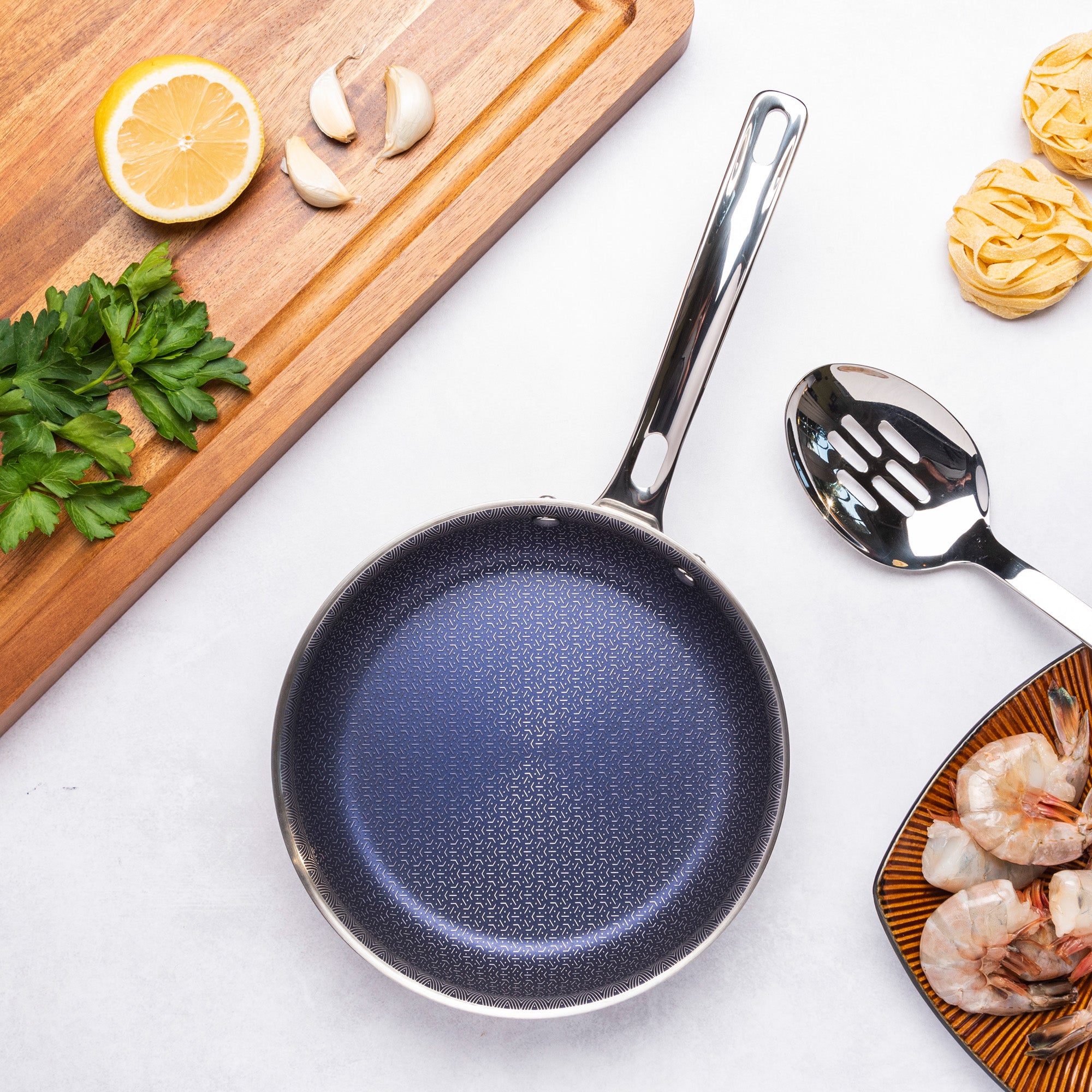 The Viking 3-Ply Hybrid Plus 8-inch Nonstick Fry Pan by Viking Culinary Products sits on a white surface, surrounded by a cutting board with parsley, garlic, and lemon, pasta nests, a slotted spoon, and a plate of raw shrimp.