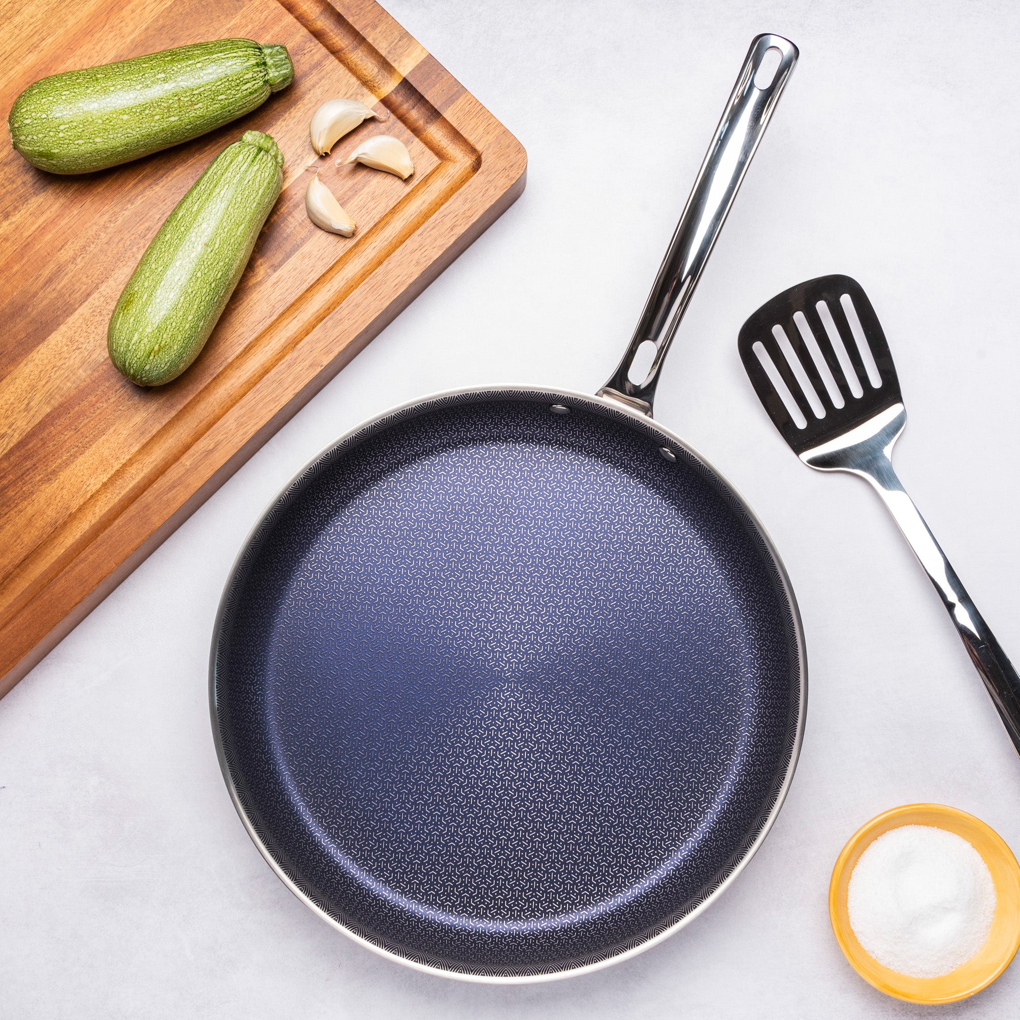 The Viking 3-Ply Hybrid Plus 12-inch Nonstick Fry Pan by Viking Culinary Products is shown with a slotted spatula, a wooden cutting board holding zucchini and garlic cloves, and a small bowl of salt on a light surface.