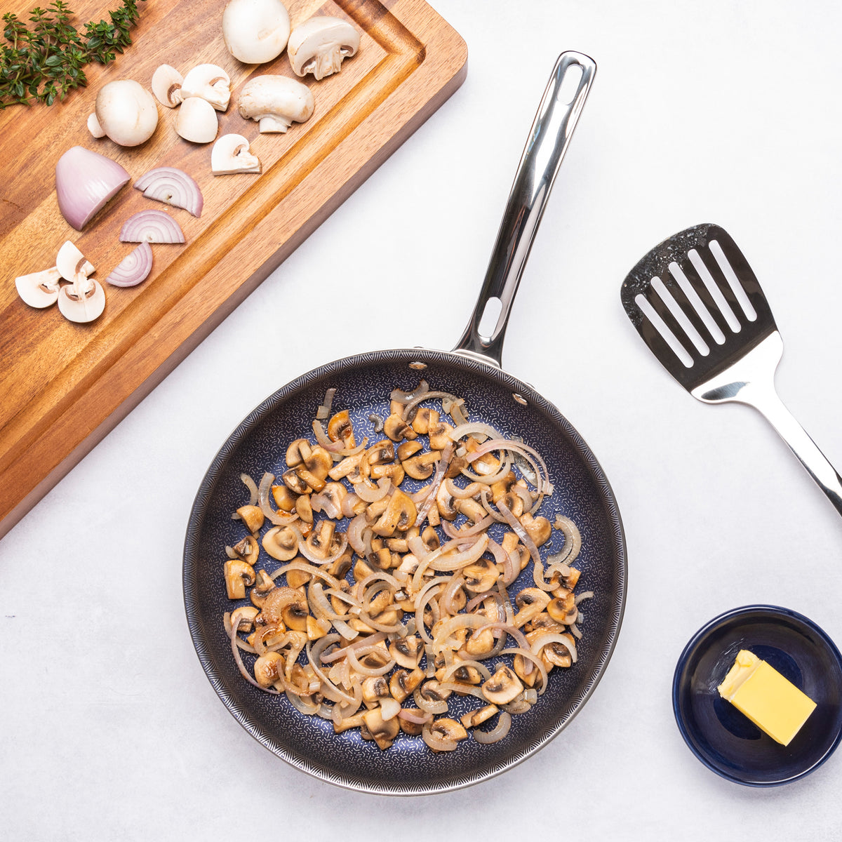 A Viking Culinary Products 3-Ply Hybrid Plus 2 Pc Nonstick Fry Pan Set (9.5" & 11") sits on a white surface next to a spatula, butter, and a wooden board with chopped mushrooms, shallots, and thyme—ideal for perfect sautéing.
