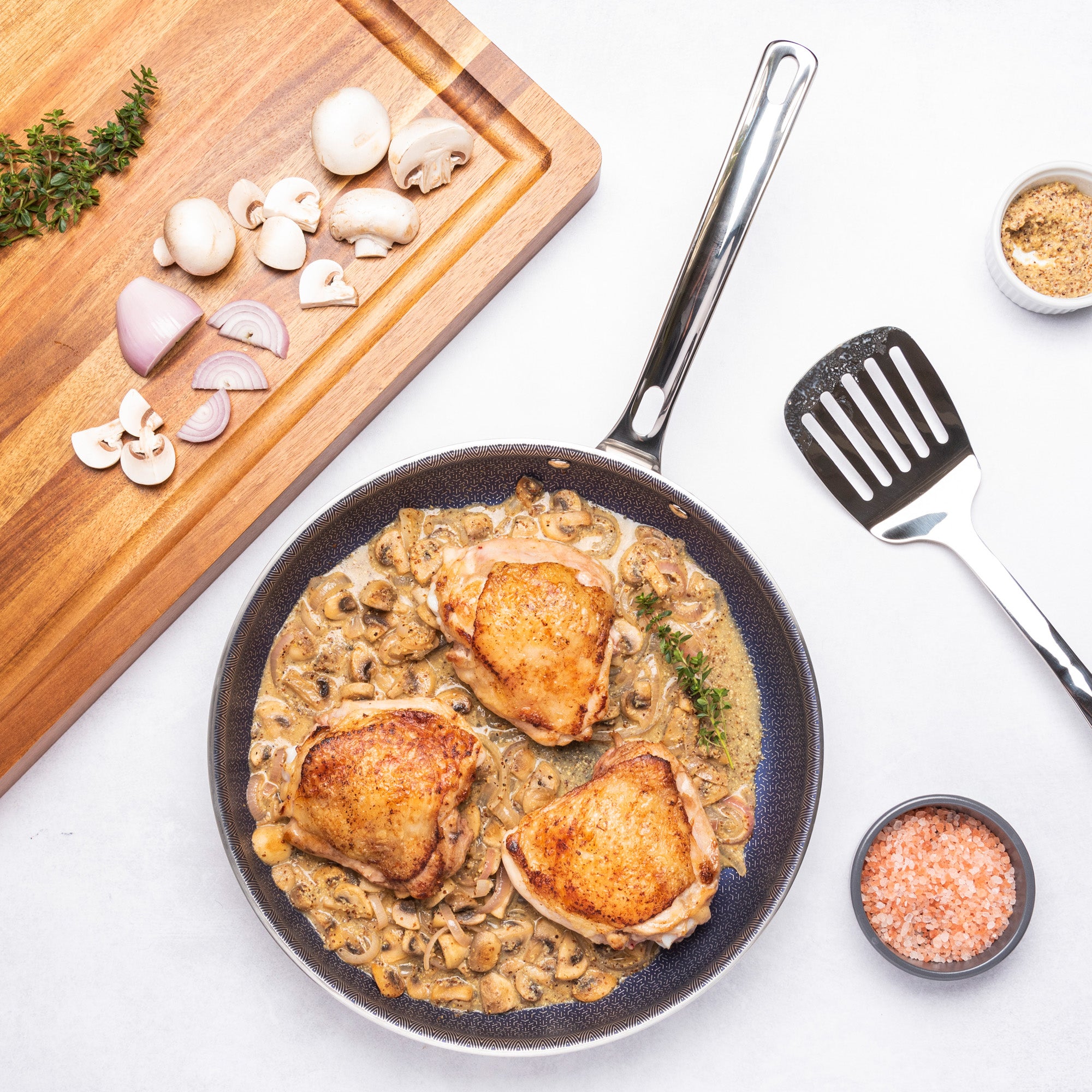 Golden-brown chicken thighs in creamy mushroom sauce cook in a Viking Culinary Products 3-Ply Hybrid Plus Nonstick Fry Pan, with sliced mushrooms, shallots, thyme, and bowls of pink salt and pepper on a nearby wooden cutting board.