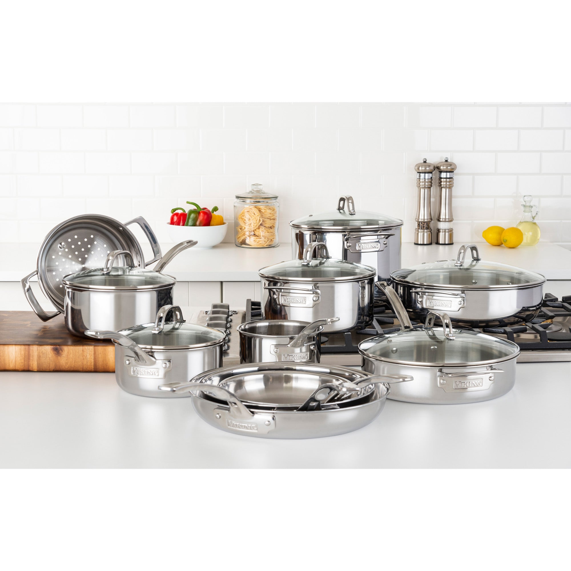 The Viking 3-Ply Stainless Steel 17-Piece Cookware Set with Glass Lids by Viking Culinary Products, including pots, fry pans, and lids, is arranged on a kitchen counter and stove with cutting boards, vegetables, pasta in a jar, pepper mills, and lemons nearby.