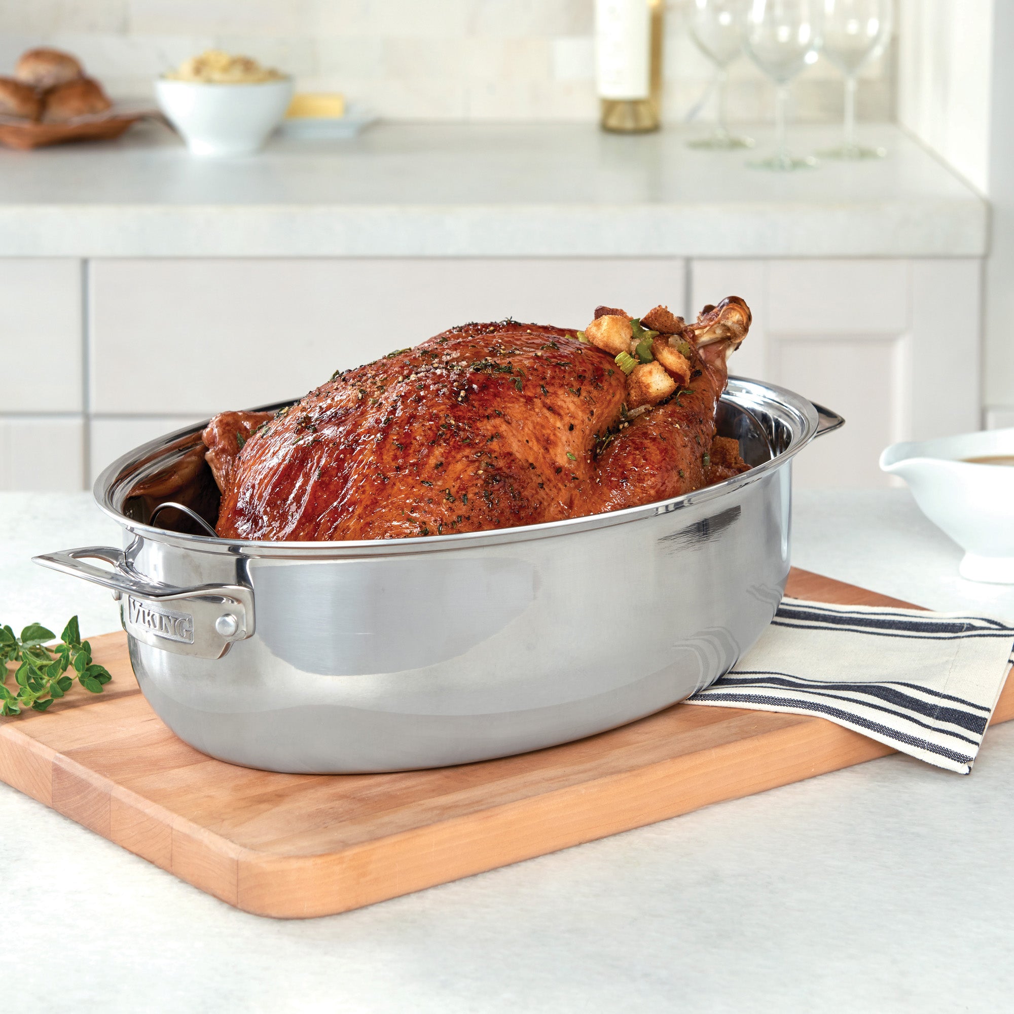 A golden-brown roasted turkey with herbs sits in the Viking 3-Ply 8.5-Quart 3-in-1 Oval Roaster with Rack by Viking Culinary Products on a wooden cutting board, beside a striped towel and an induction lid on the kitchen counter.