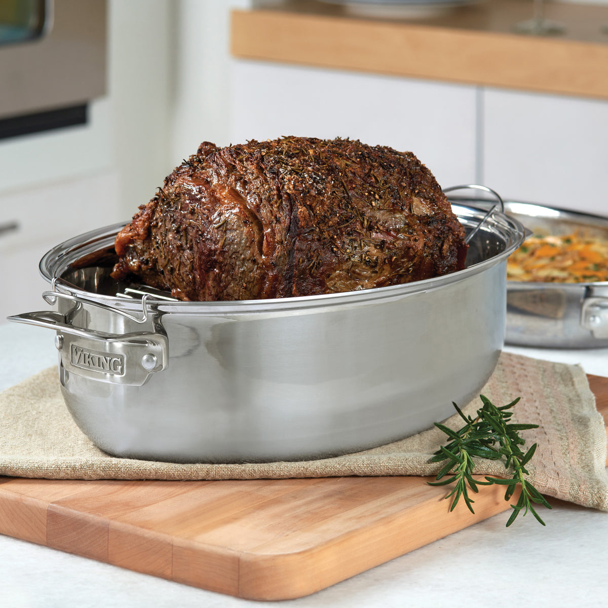 A cooked, seasoned roast sits in the Viking 3-Ply 8.5-Quart 3-in-1 Oval Roaster with Rack by Viking Culinary Products on a kitchen counter, accented with rosemary, showcasing the quality of this stainless steel cookware.