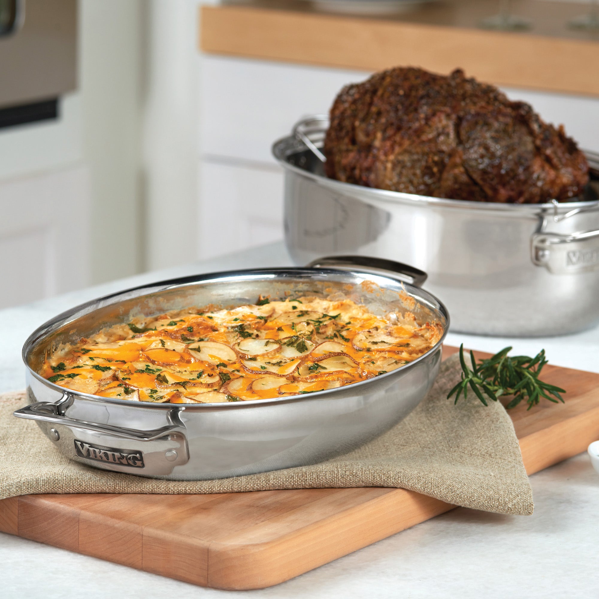 The Viking 3-Ply 8.5-Quart 3-in-1 Oval Roaster with Rack by Viking Culinary Products holds roasted meat, while cheesy scalloped potatoes sit nearby on a wooden cutting board with fresh rosemary in the background.