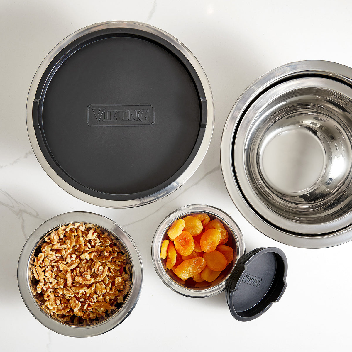 The Viking 10-Piece Stainless Steel Mixing Bowl Set with Lids by Viking Culinary Products, shown on a white countertop, includes bowls filled with walnuts and dried apricots and others empty—all versatile enough to double as mixing bowls.