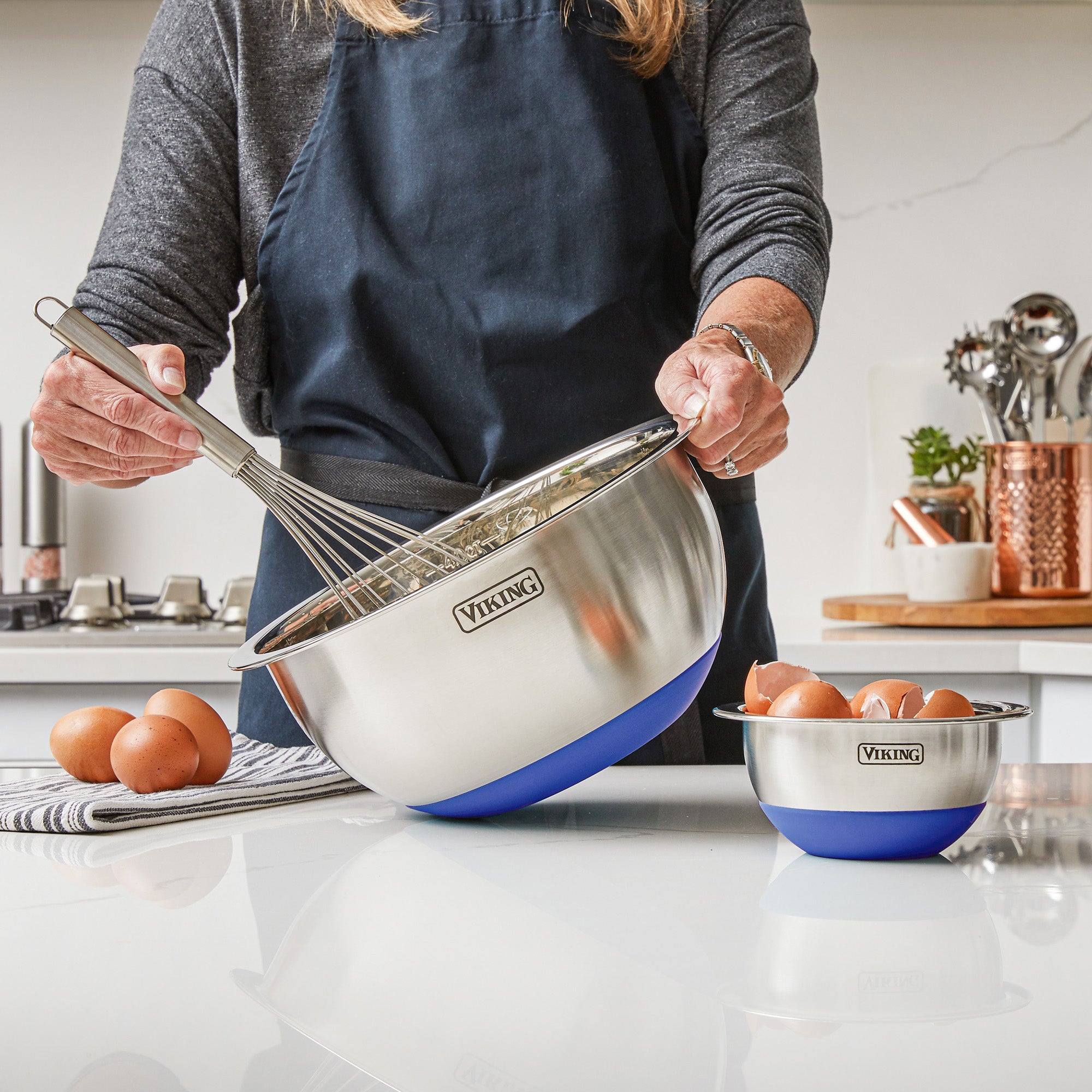 Wearing a navy apron, someone whisks ingredients in a large blue Viking Culinary Products mixing bowl from the 10-piece stainless steel set with lids, while eggs and a smaller Viking bowl sit nearby. Kitchen utensils and copper containers are in the background.