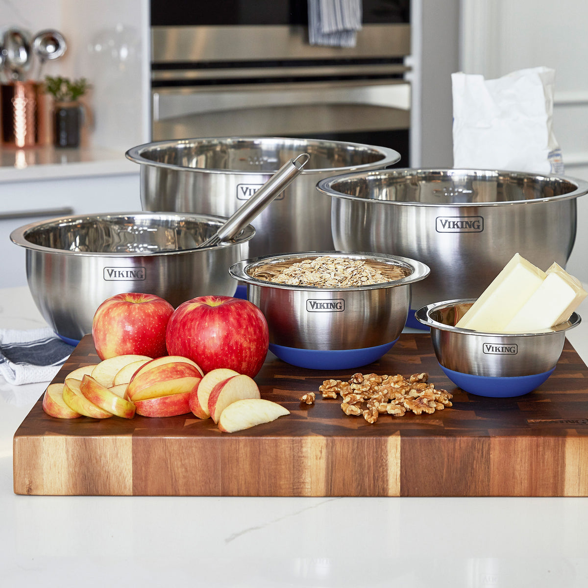 The Viking Culinary Products 10-Piece Stainless Steel Mixing Bowl Set with Lids in Blue sits on a wooden board in a modern kitchen alongside apples, apple slices, walnuts, oats, butter, and a bag of flour.