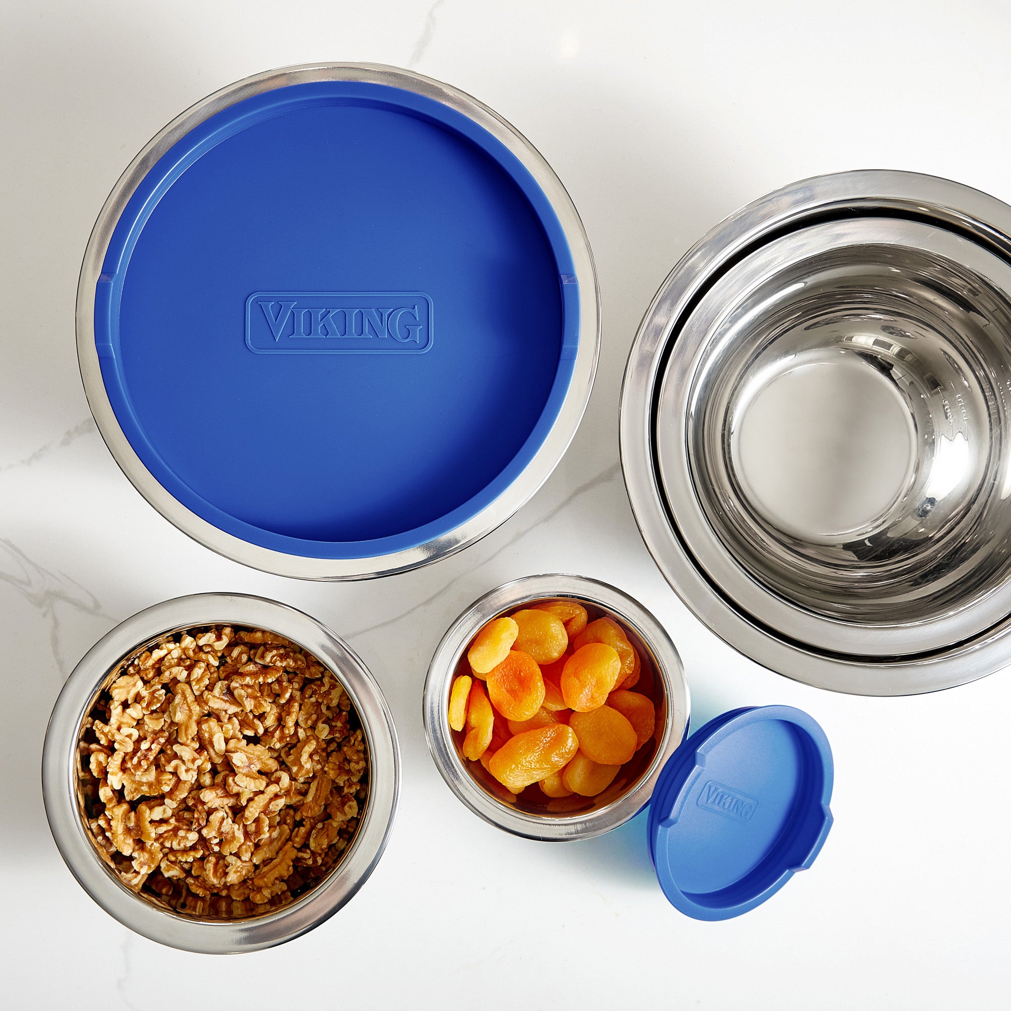 Four Viking Culinary Products mixing bowls from the 10-piece stainless steel set with blue lids are shown; two hold dried apricots and walnuts, while the others are empty, all arranged on a white surface.